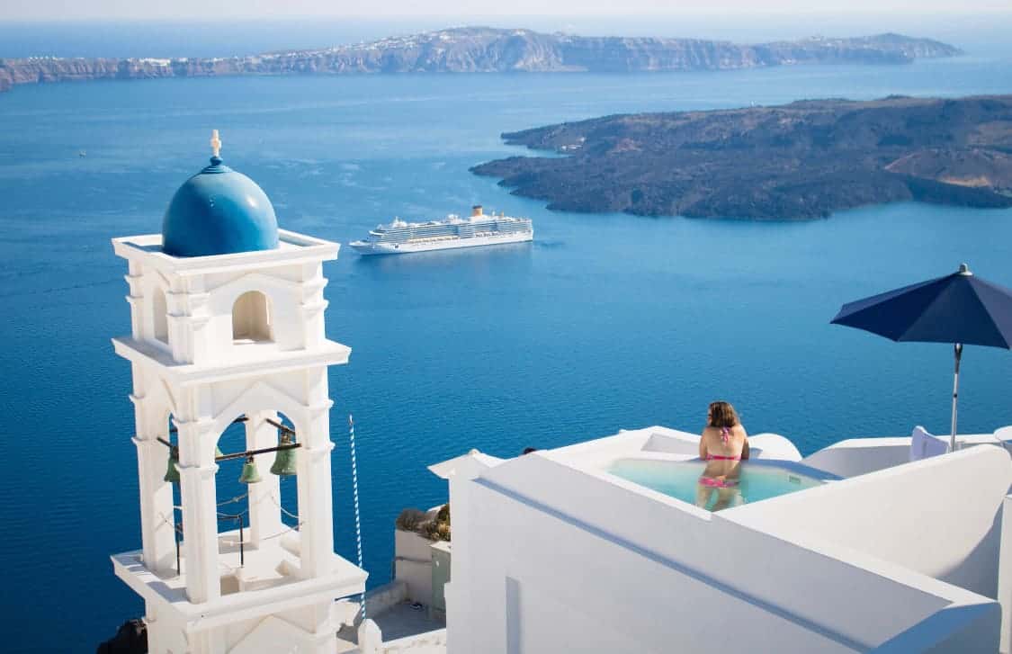 White building with a rooftop pool with a female in a bikini in the pool over looking the water in Greece where there is a cruise ship going by