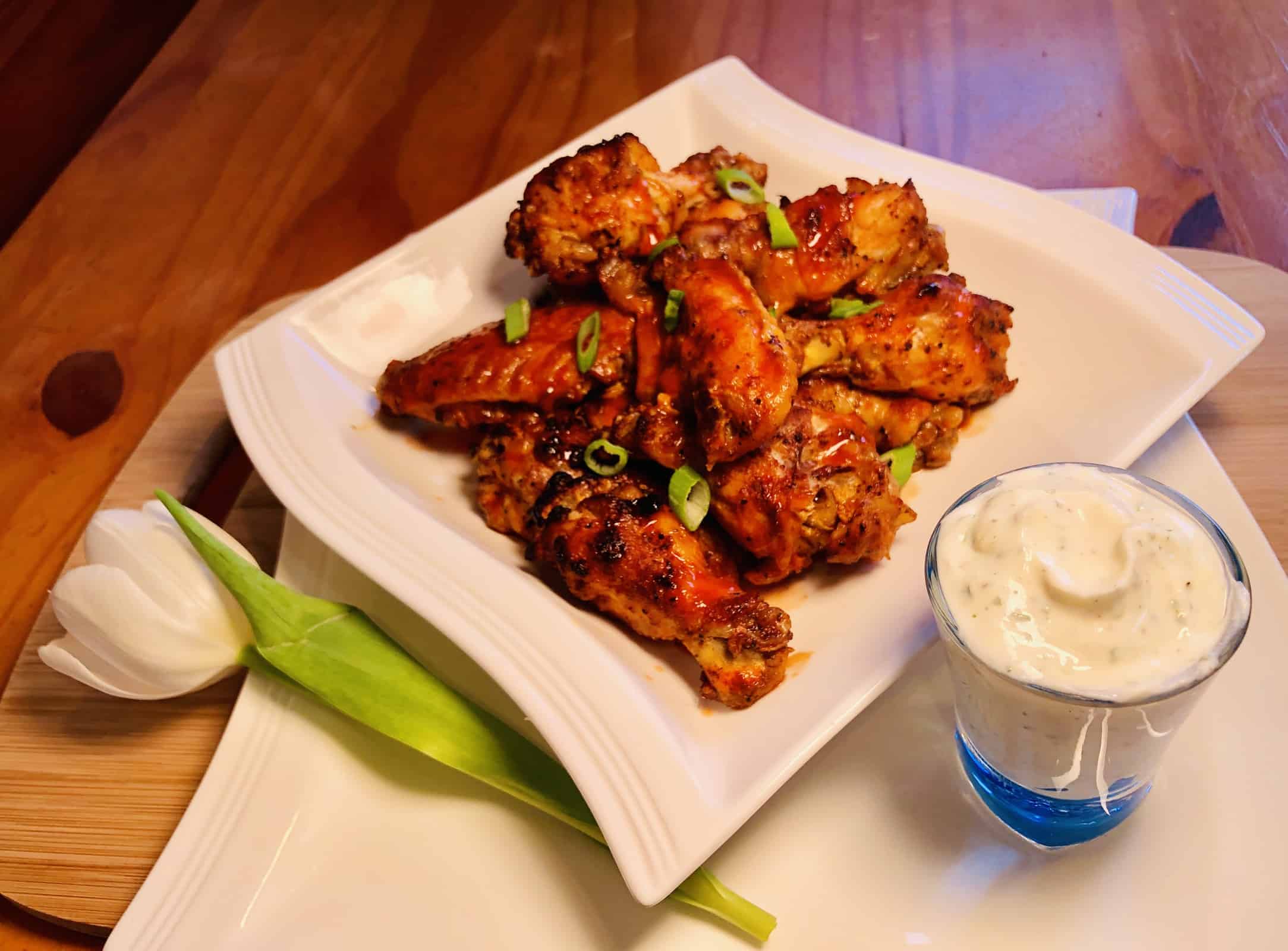Keto Buffalo Chicken Wings with chives on a white plate with ranch dressing in a shot glass and a white tulip on a wooden table