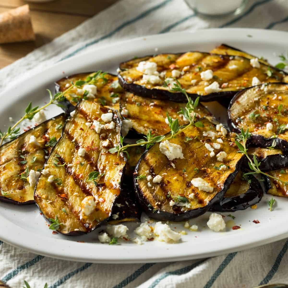 Grilled eggplant slices topped with feta cheese on a white plate on a white and green striped placemat