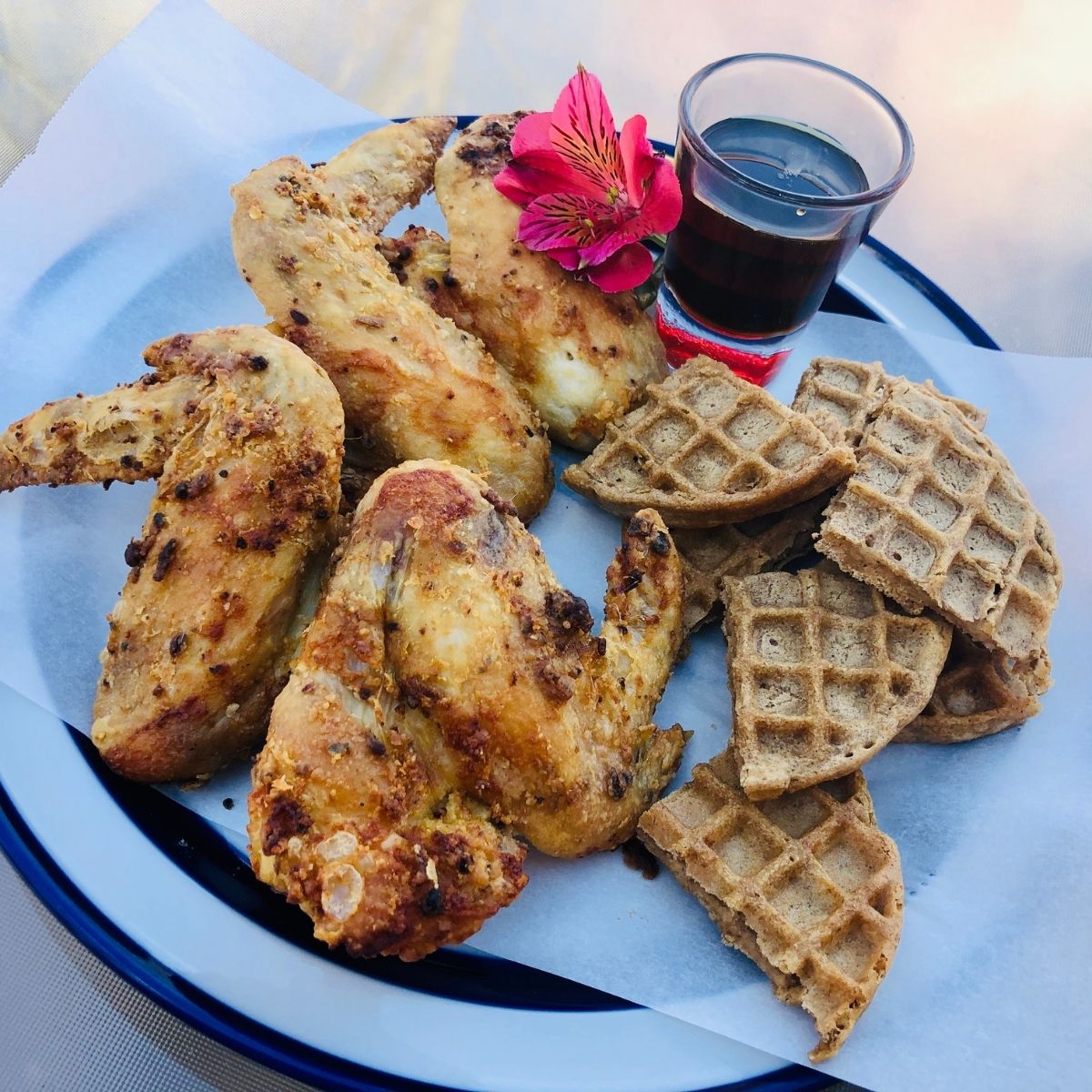Air Fried chicken wings and waffles on a sheet of parchment paper on a plate with a side of syrup and a pink flower