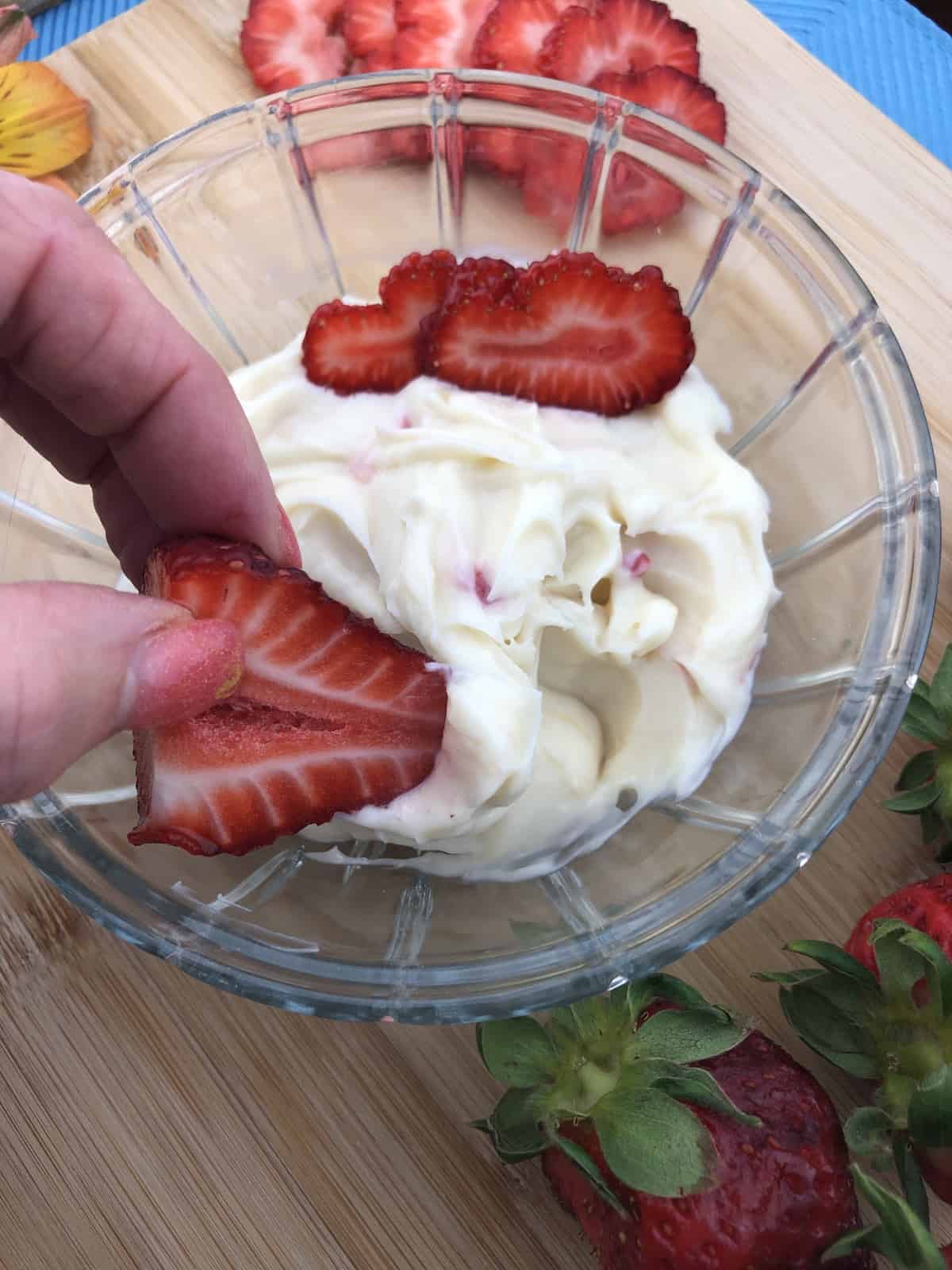 Dipping Strawberries in cream cheese dip from a glass bowl sitting on a wooden cutting board