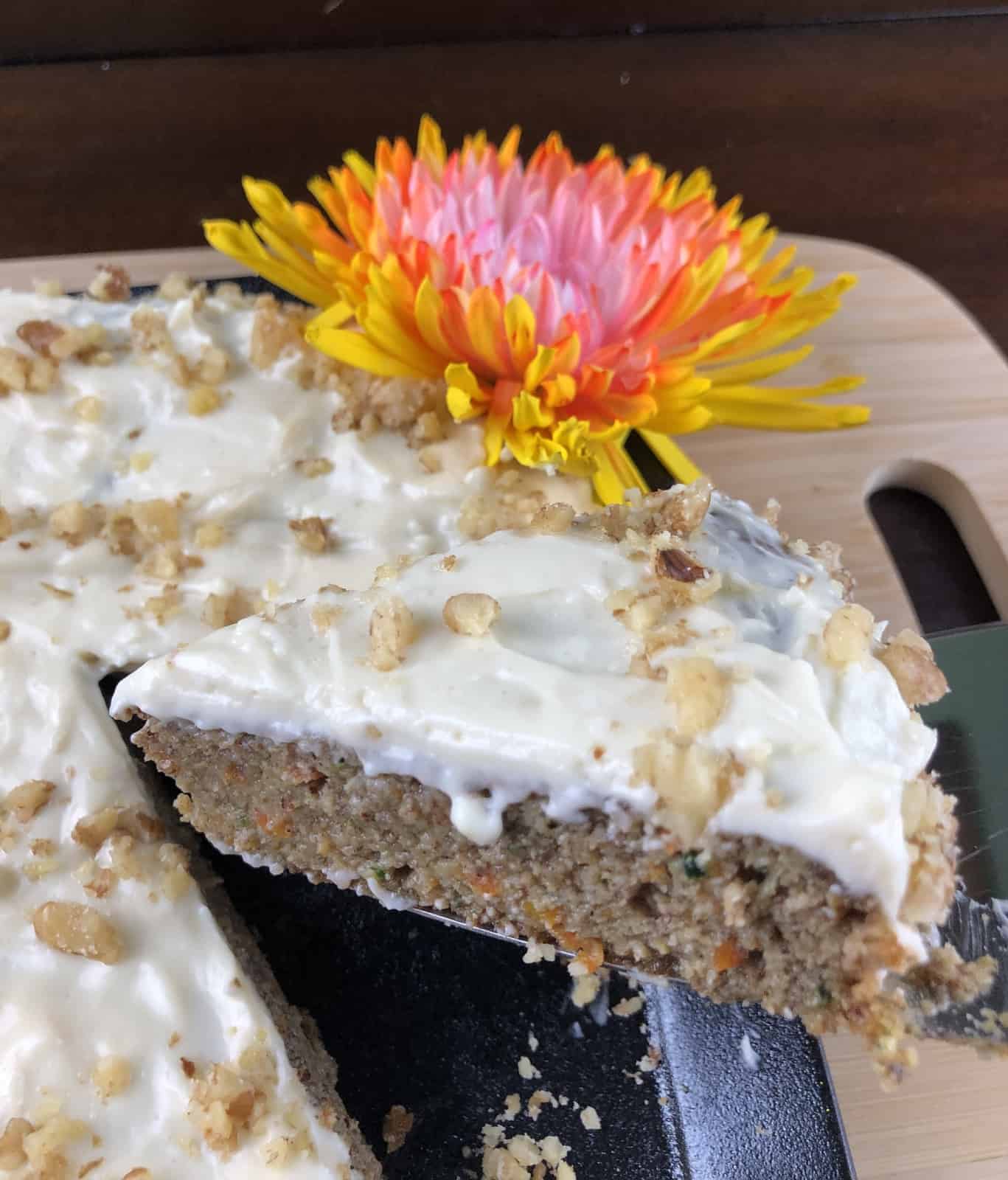 Carrot Cake with Cream Cheese Frosting sprinkled with walnuts on a wooden cutting board with a yellow and pink flower