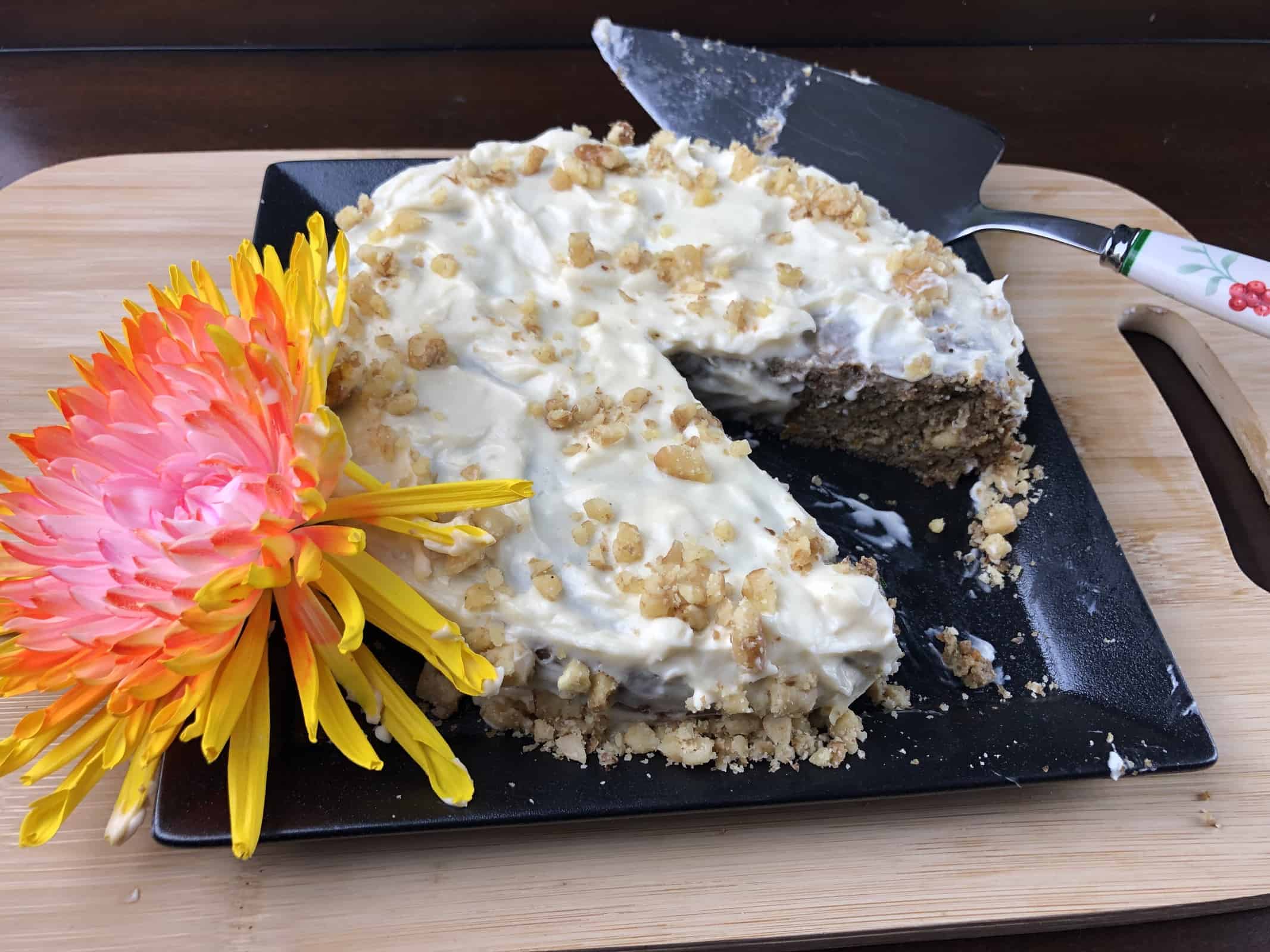 Keto Carrot Cake with cram cheese frosting and walnuts sprinkled on top on a black plate with a flower and a cake server on a wooden cutting board