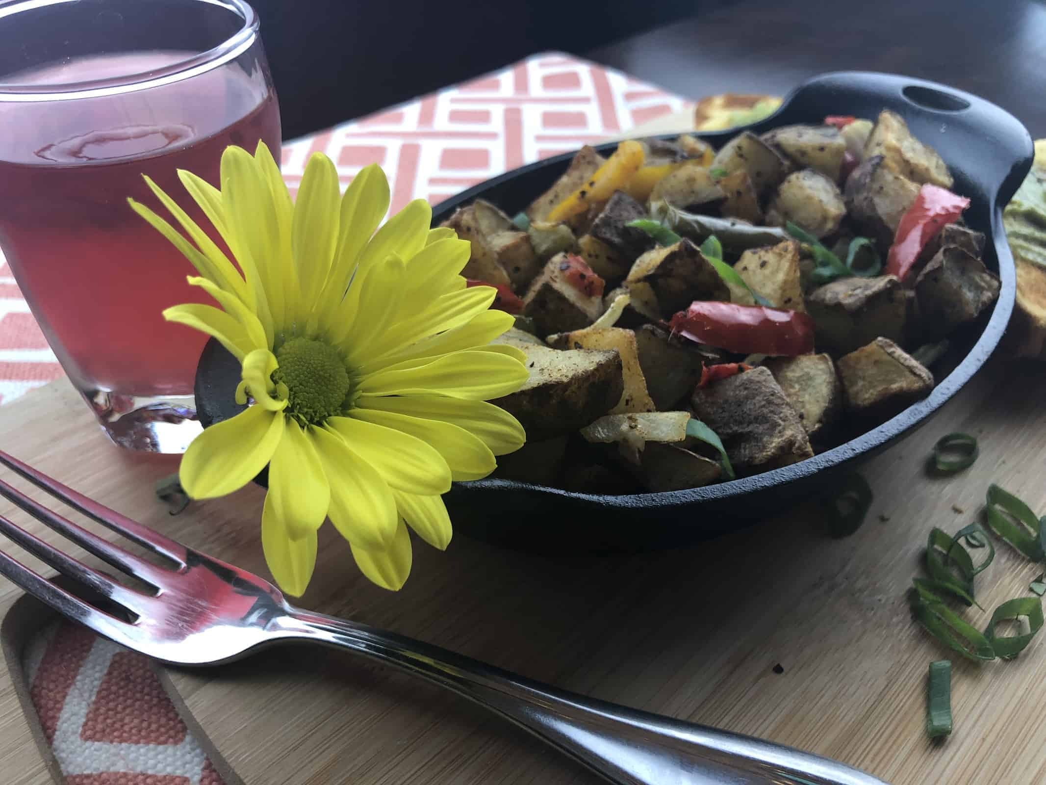 Air Fried Home Fries with peppers in a black casserole dish on a wooden cutting board with a yellow flower and a glass of fruit punch