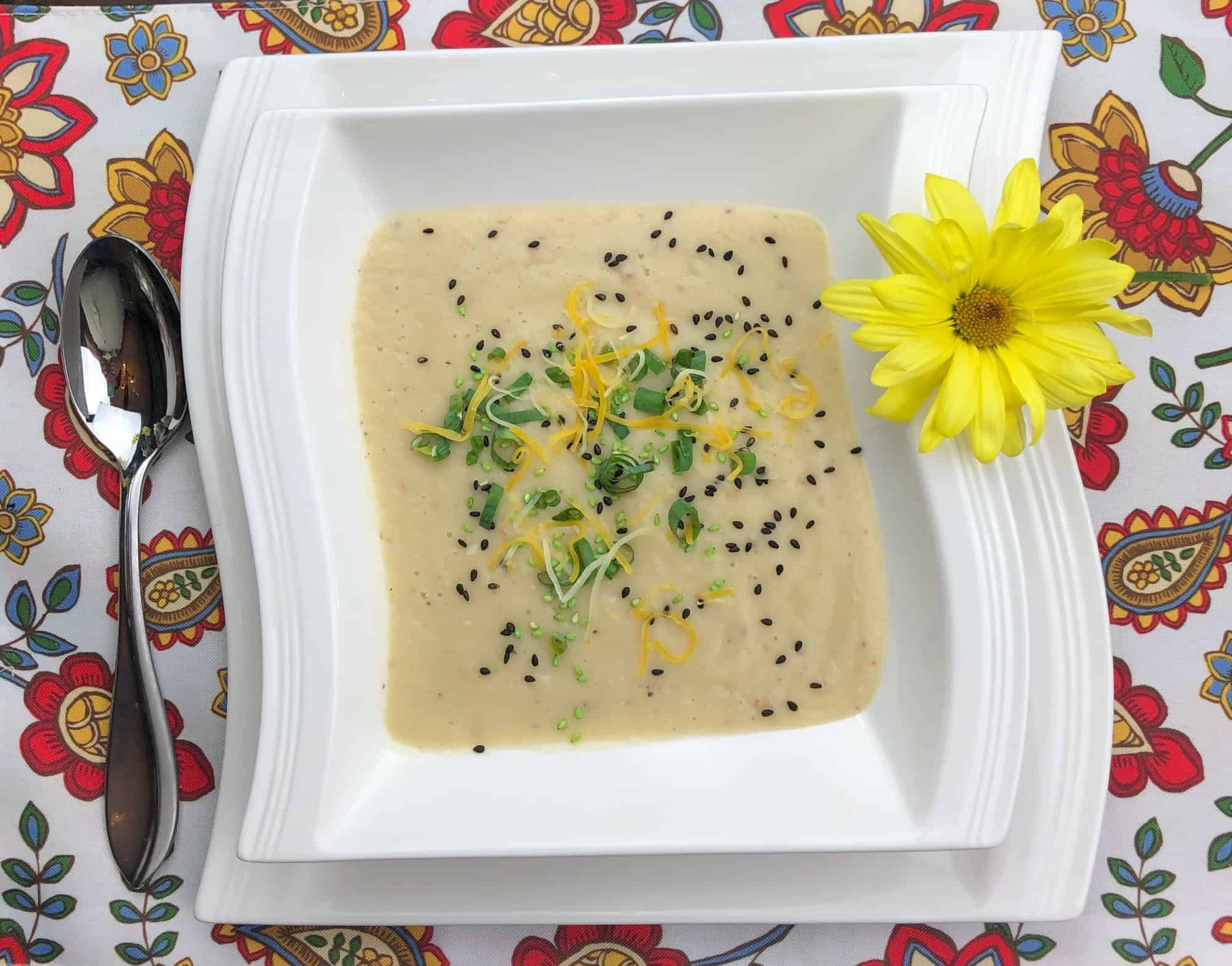 Cauliflower Bisque with chives shredded cheese and black sesame seeds in a white bowl with a yellow flower and a spoon on a printed tablecloth