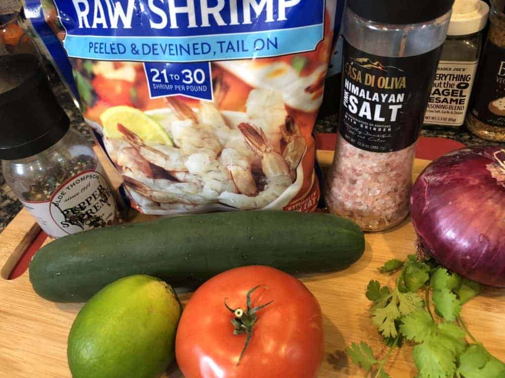  Ingredients for shrimp ceviche displayed on a wooden cutting board - Pepper, Bag of frozen shrimp, salt, cucumber, onion, parsley, lime and a tomato