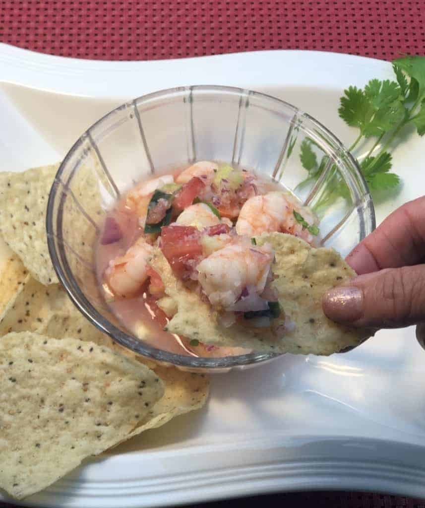 Scooping shrimp ceviche onto a Tostito corn chip from a clear glass bowl filled with shrimp ceviche sitting on a white plate