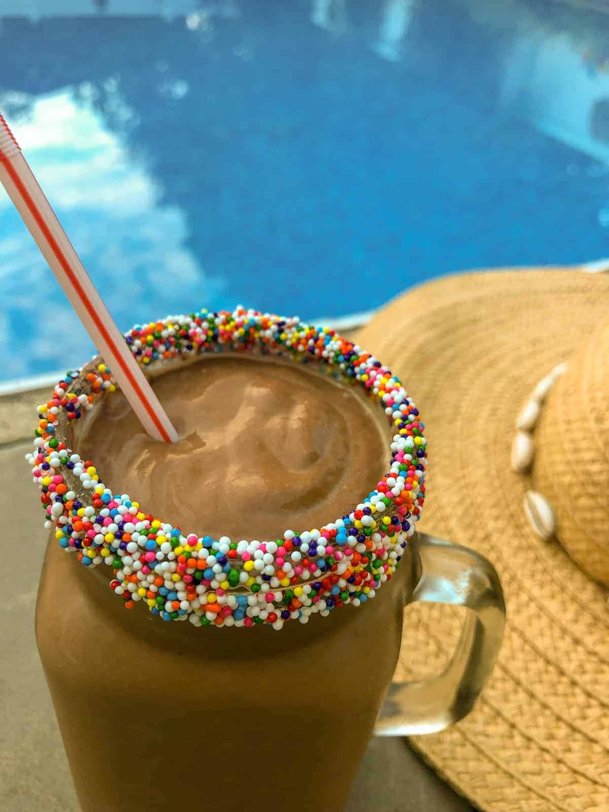 Chocolate Avocado Milkshake in a clear glass jar mug with colored sprinkles on the rim by the pool with a straw hat