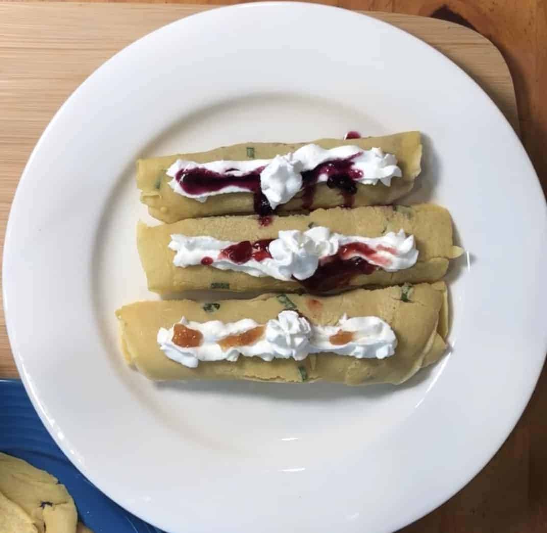 3 crepe rollups with whipped cream and jelly on a white plate on a wooden cutting board