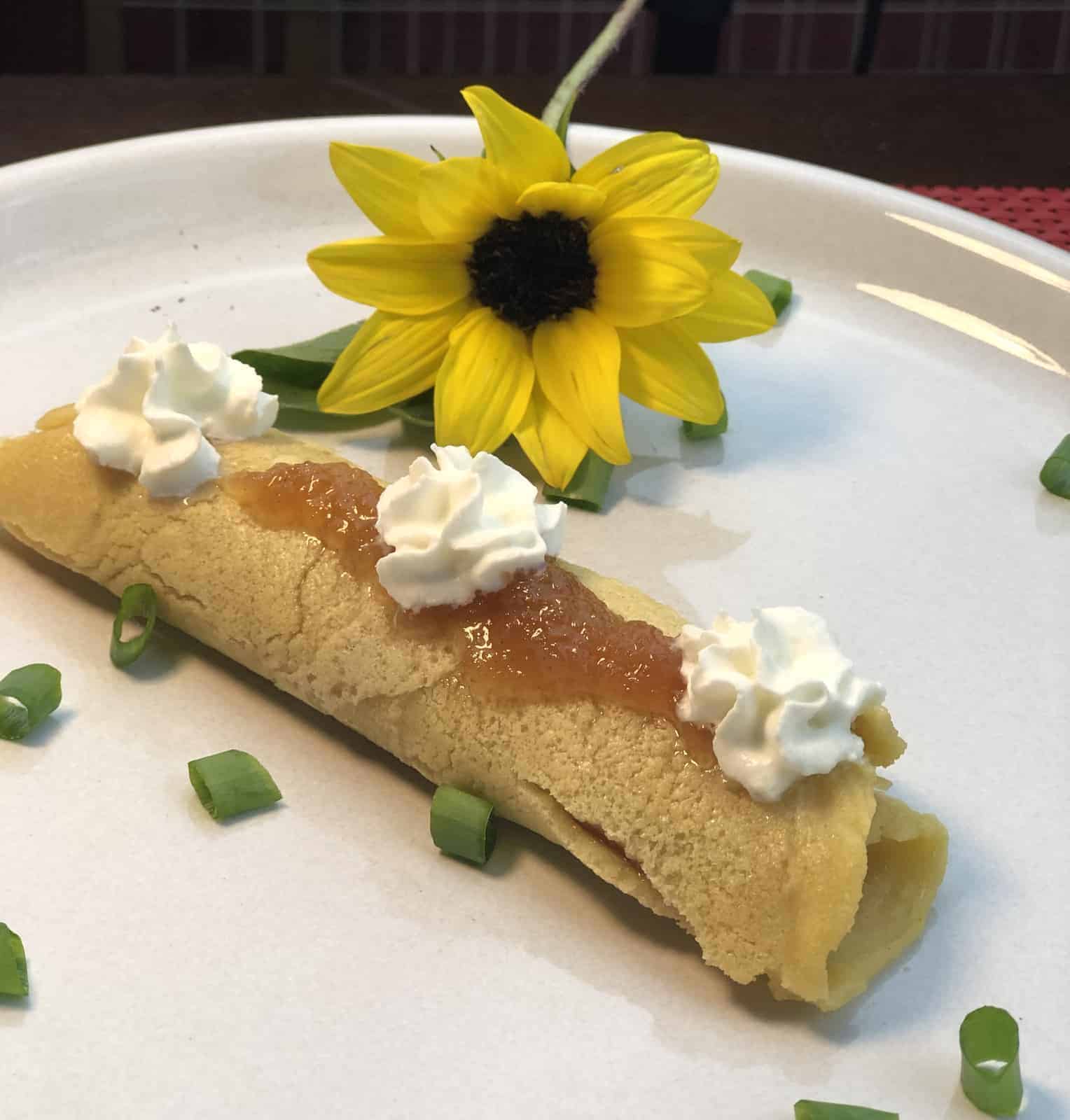  Chickpea Crepe with whipped cream and jelly on top sitting on a white plate with chives and a yellow flower