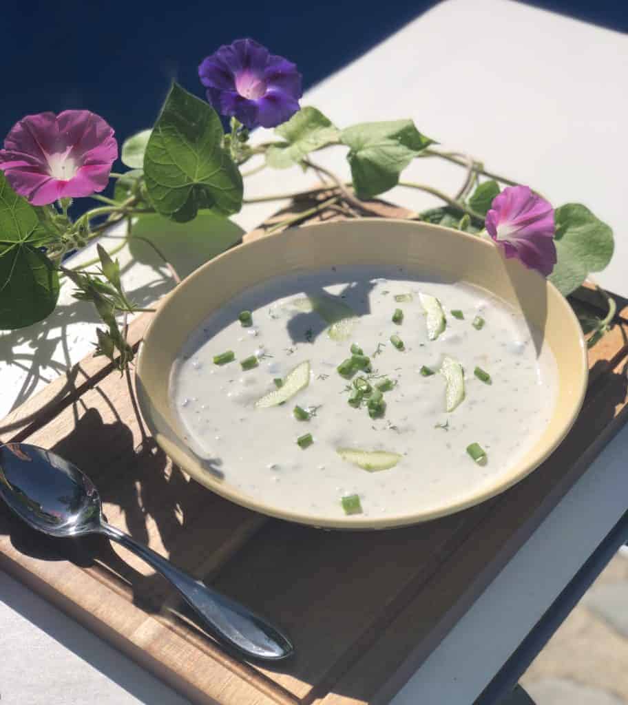 chilled cucumber soup in a yellow bowl on a wooden cutting board with a spoon and purple and pink morning glory flowers
