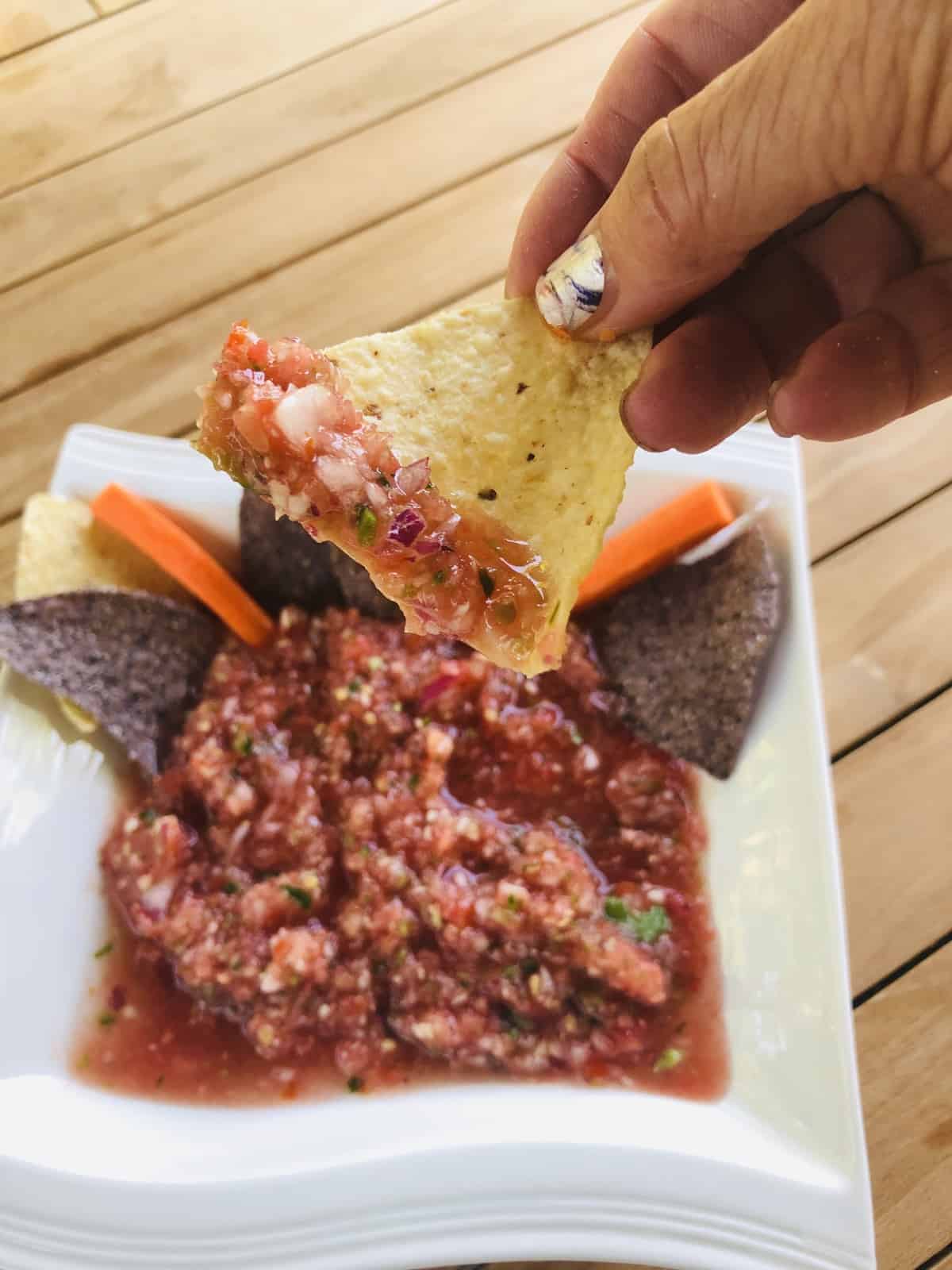 Dipping a yellow Tostito chip in a bowl salsa sitting on a wooden table