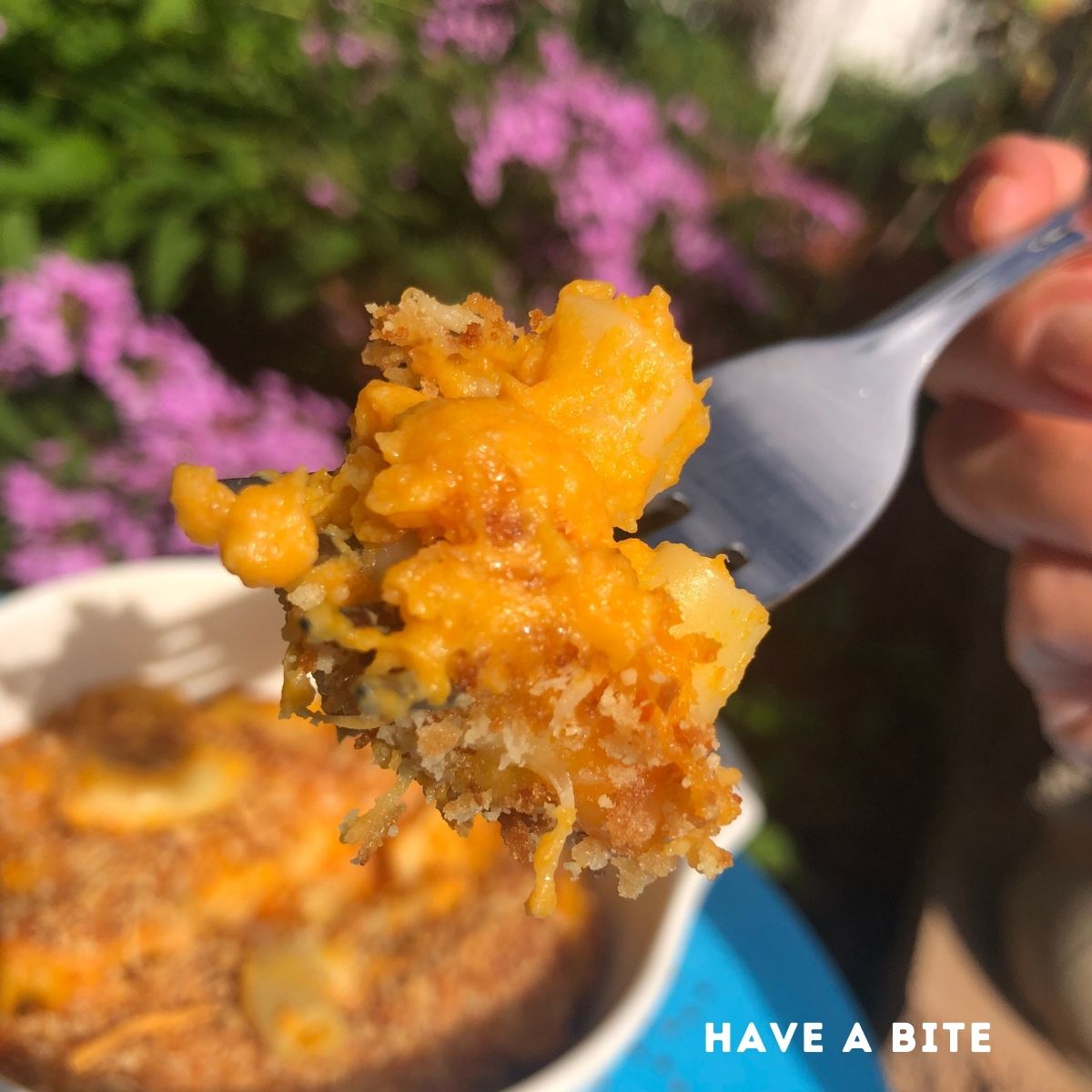 a forkful of baked macaroni and cheese over a bowl of Mac and cheese on a blue table in a garden with purple flowers