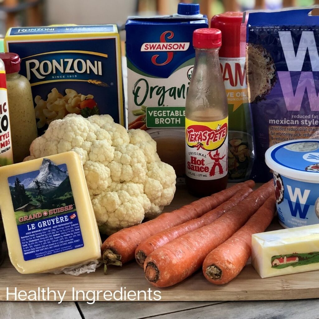 Panko bread crumbs, pepper,a block of Gruyere cheese, cauliflower, carrots, Ronzoni macaroni, vegetable broth, hot sauce, WW cheese, WW cream cheese and salt displayed ingredients on a wooden cutting board for Mac and cheese recipe