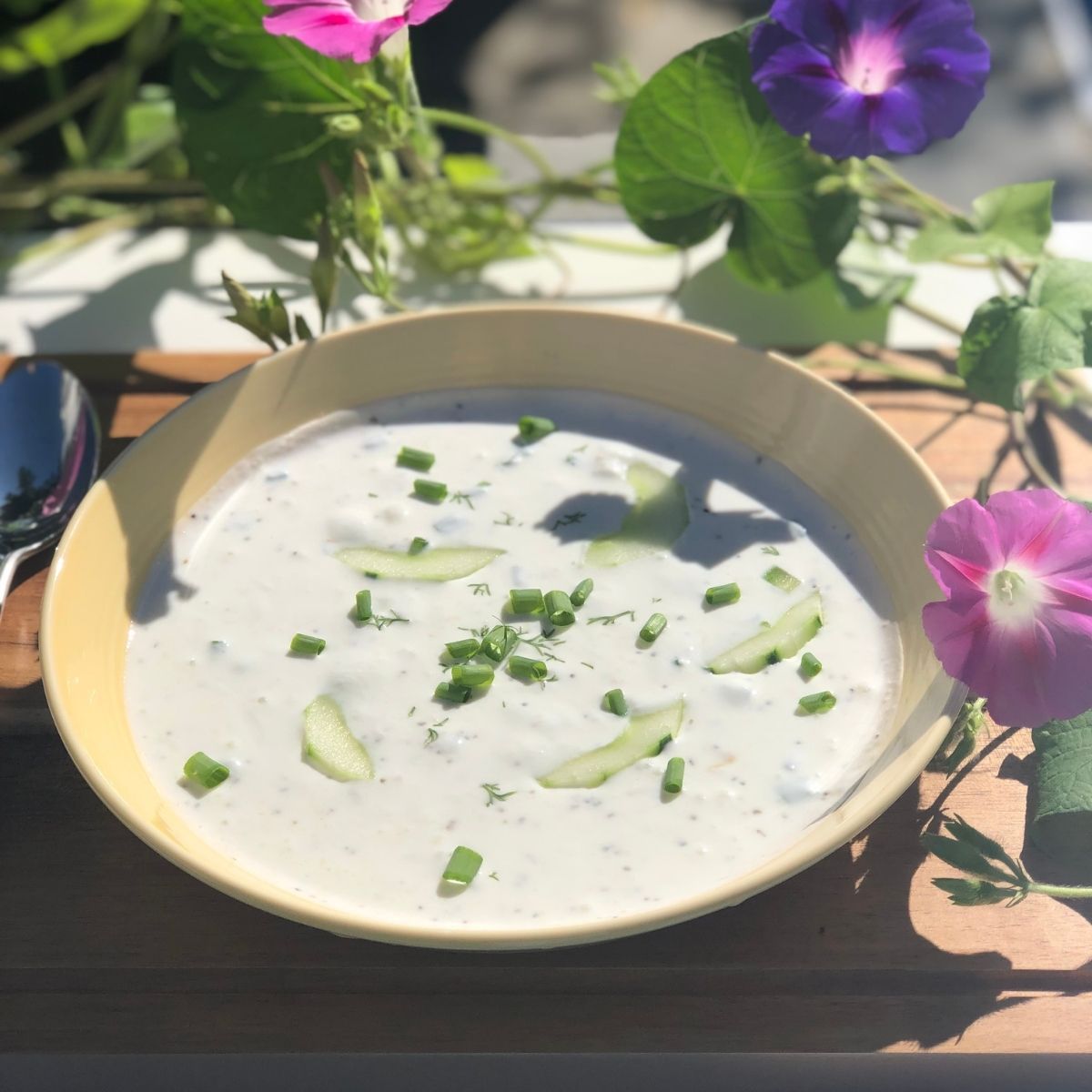 Chilled keto cucumber soup in a yellow bowl on a wooden table in a garden of pink and purple morning glories