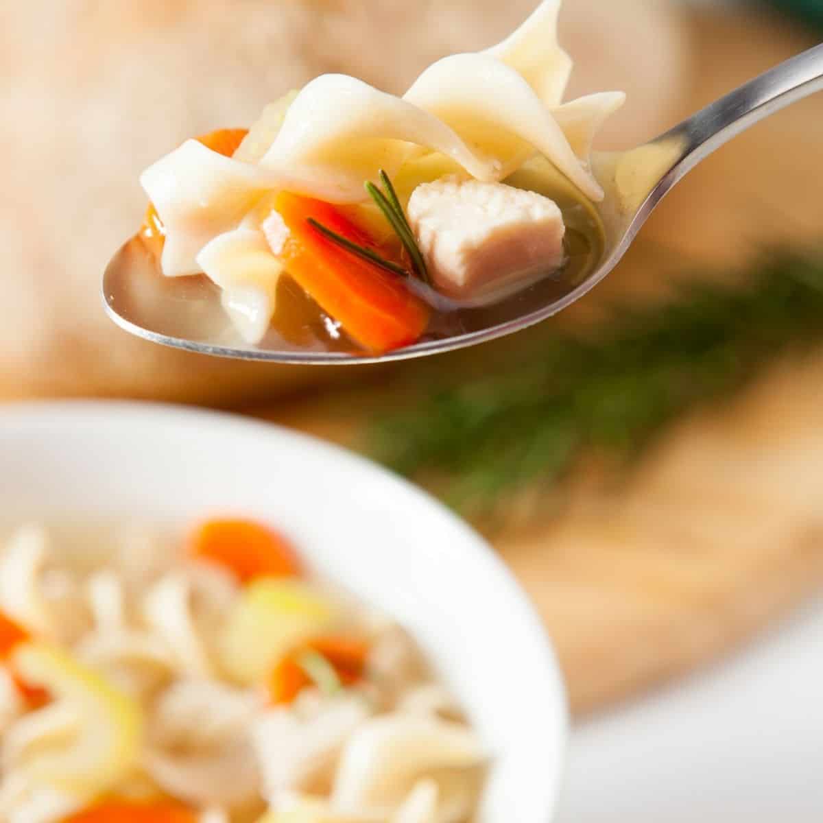 A bowl of chicken noodle soup with a spoon with a serving of noodles chicken and carrots