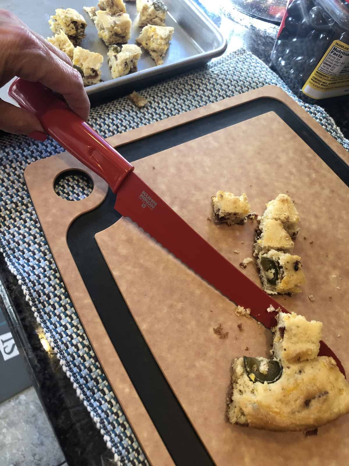 Cutting keto cornbread into cubes for stuffing with a red knife on a cutting board and a silver baking sheet  with bread cubes on it