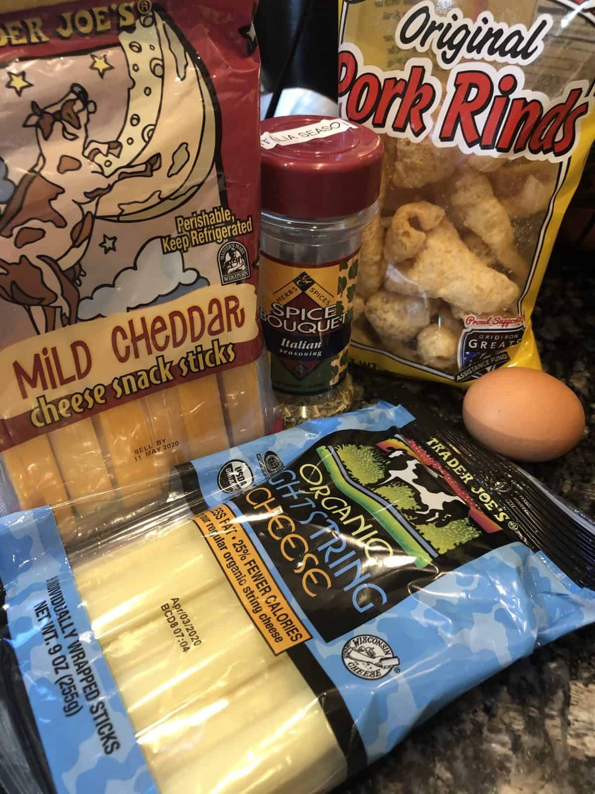 Ingredients for Low Carb Mozzarella sticks which includes a package of mild cheddar string cheese and Trader Joes Mozzarella string cheese and a bag of pork rinds and one brown egg and Italian seasoning