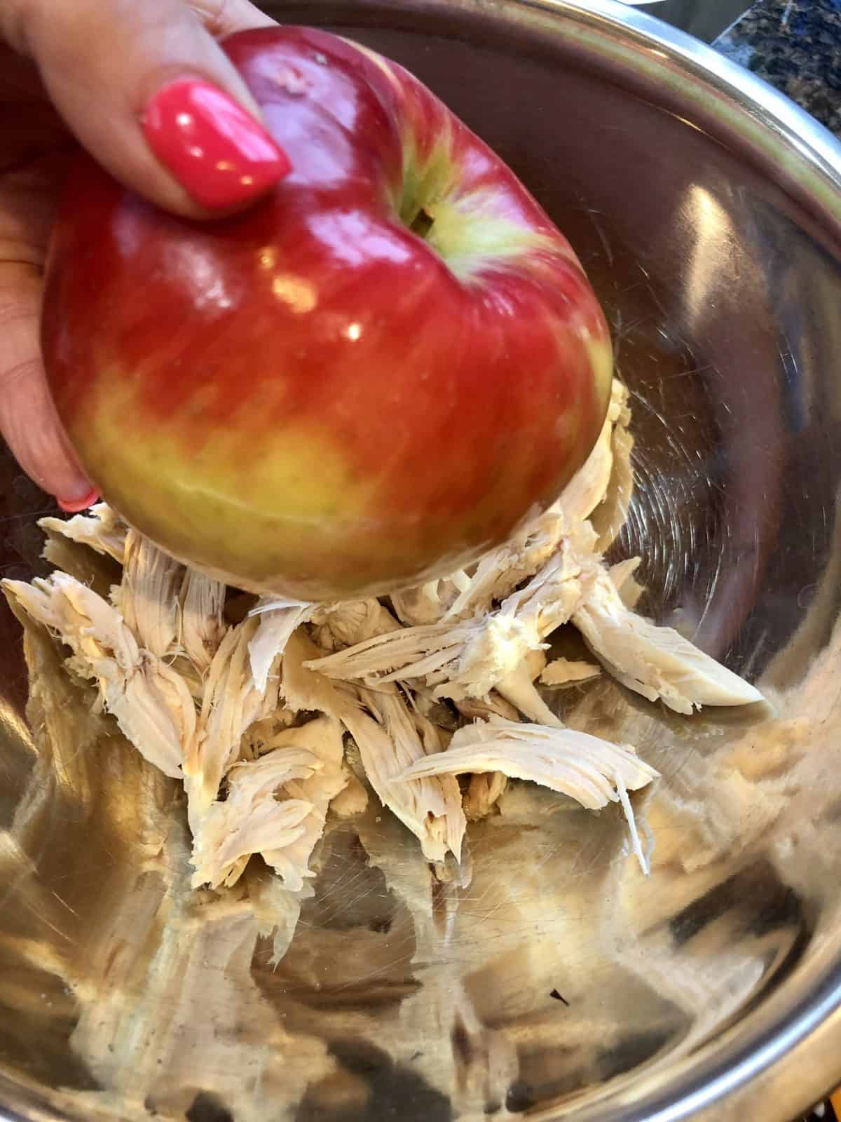 A hand holding a Honeycrisp Apple over a bowl of shredded Chicken