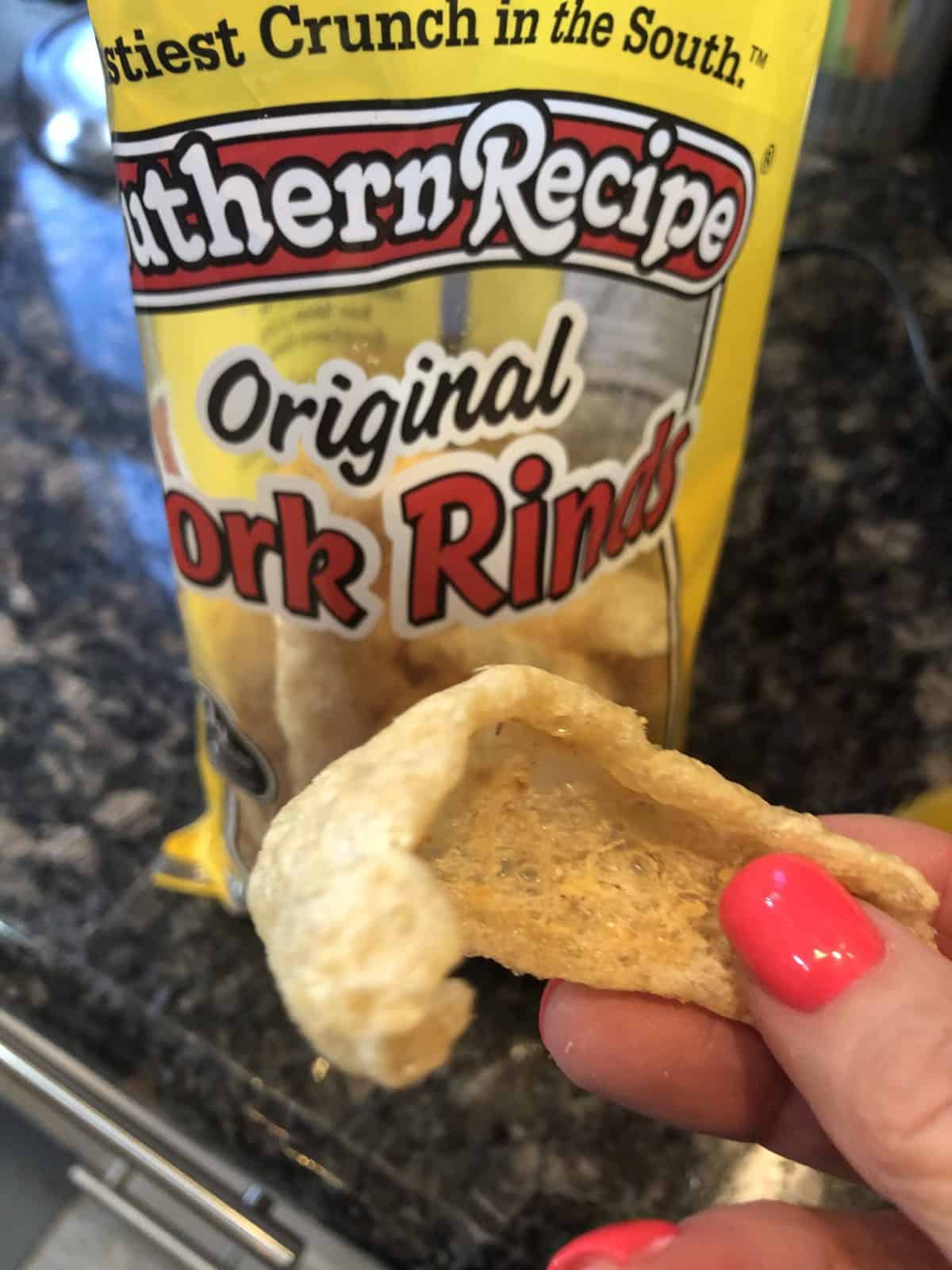 A hand holding a Pork Rind in front of a bag of Southern Recipe Original Pork Rinds