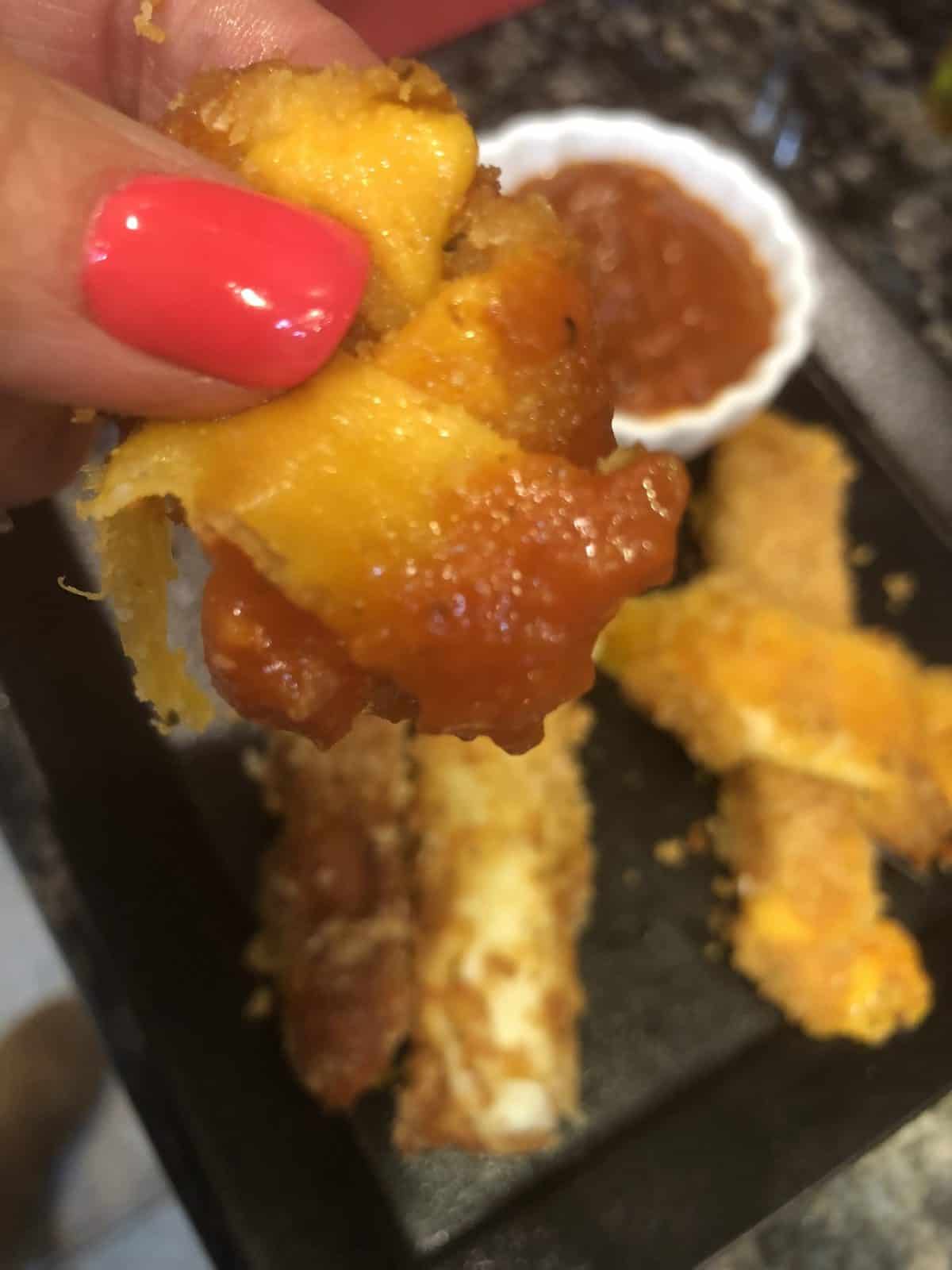 A hand holding a Dipped breaded cheese stick in tomato sauce