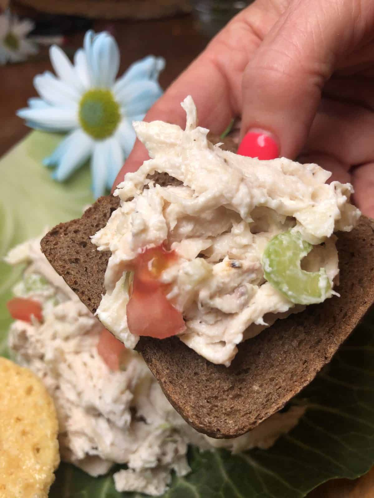 Chicken Salad on Pumpernickel Bread being held by a hand over a plate of homemade rotisserie chicken salad