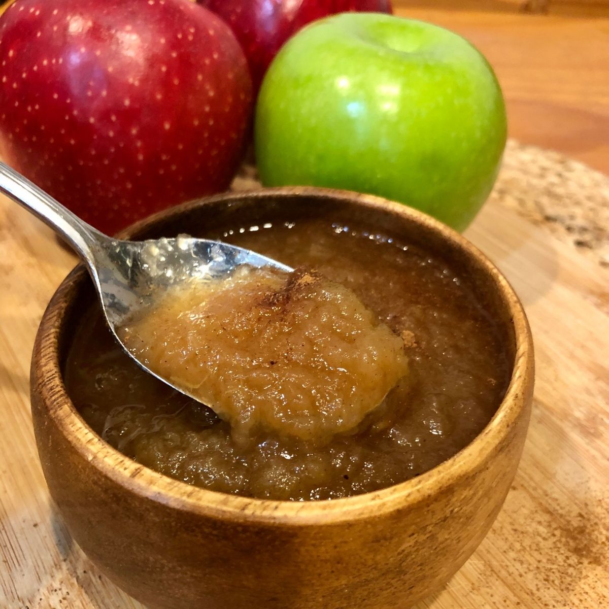 Unsweetened applesauce in a wooden bowl with a spoonful of applesauce with a red and green apple on the side