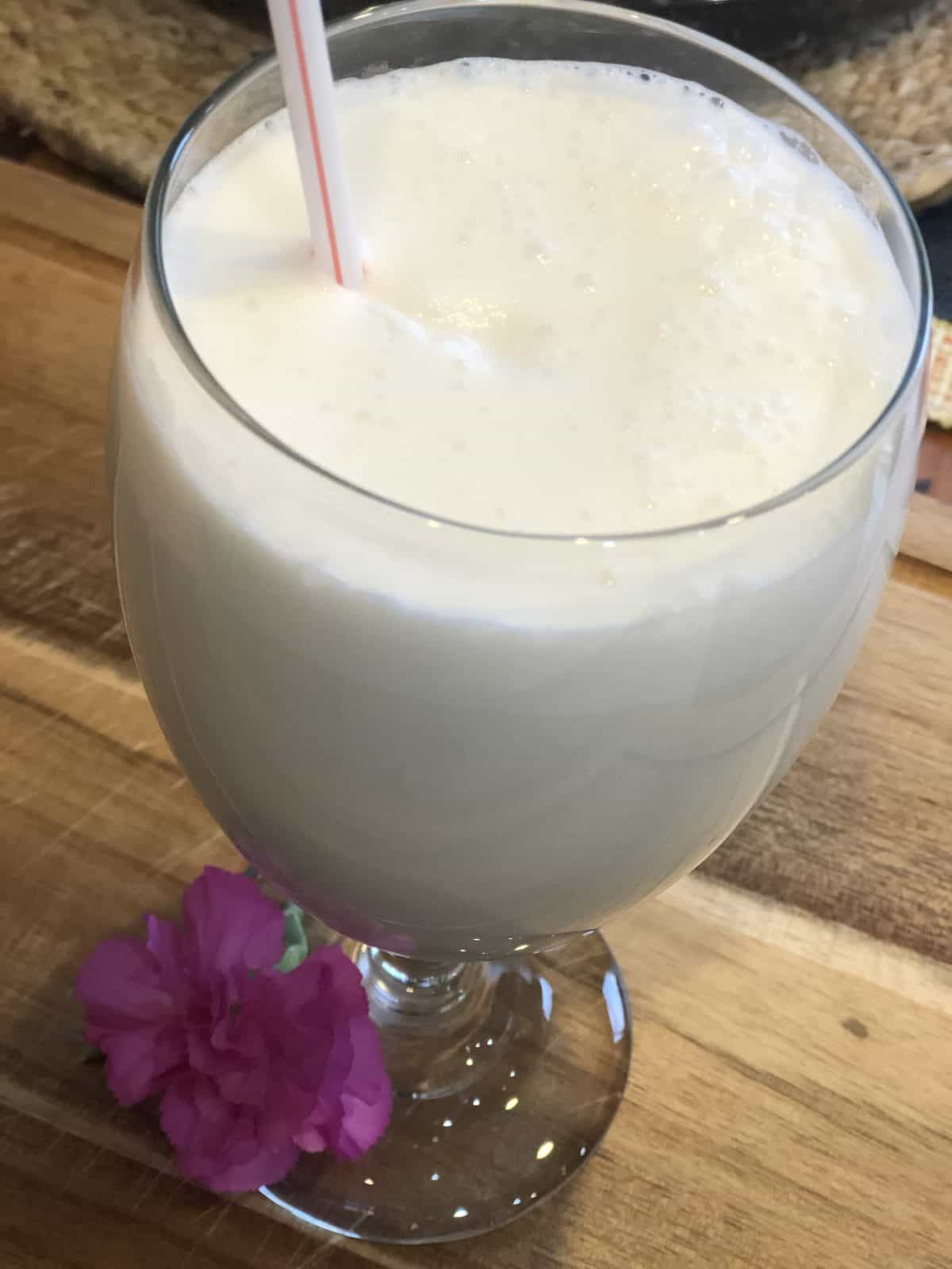 Low Carb Vanilla Frosty in a clear glass with a straw and a pink flower on a wooden cutting board