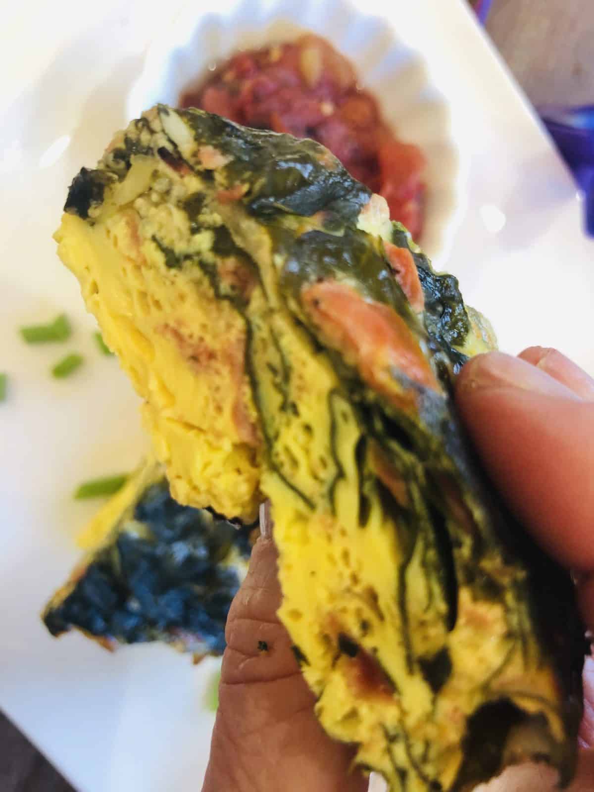 A slice of Spinach Frittata held in a hand over a white plate with green scallions and salsa