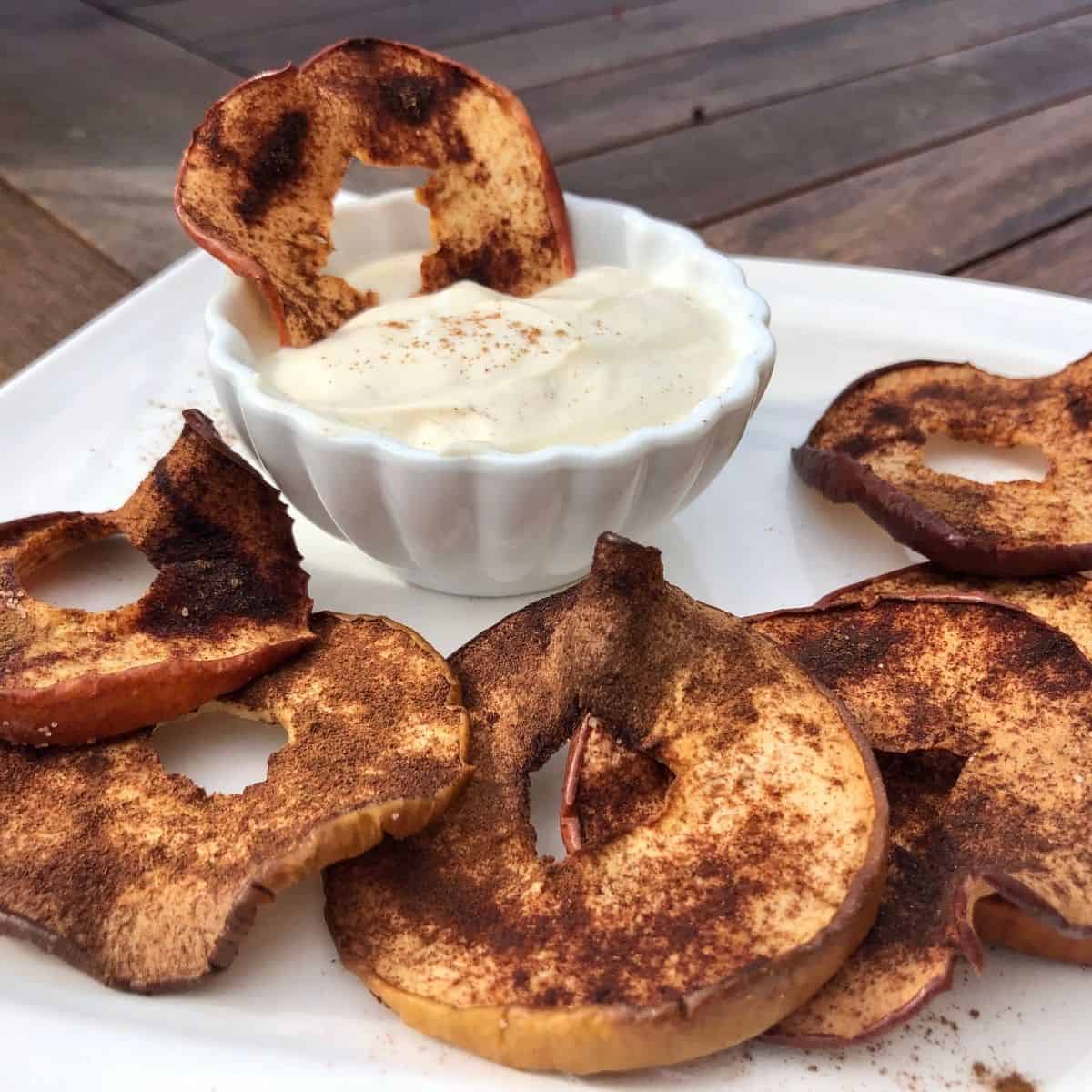 Apple chips on a white plate and an apple chip being dipped in a cream cheese dip in a small white bowl sitting on the plate on a wooden table