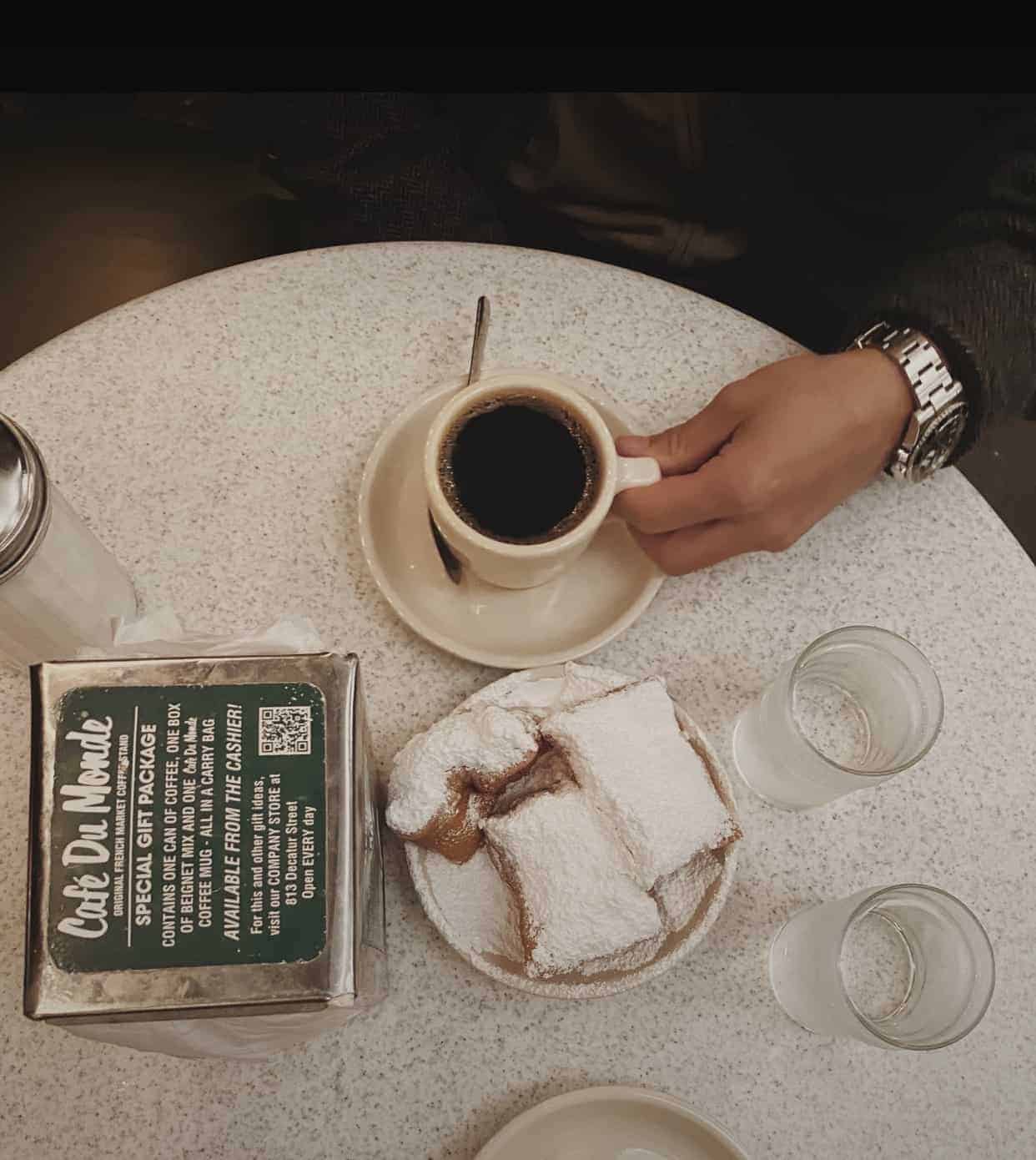 A hand holding the handle of a coffee cup with coffee init on a white table with beignets at Cafe Du Monde