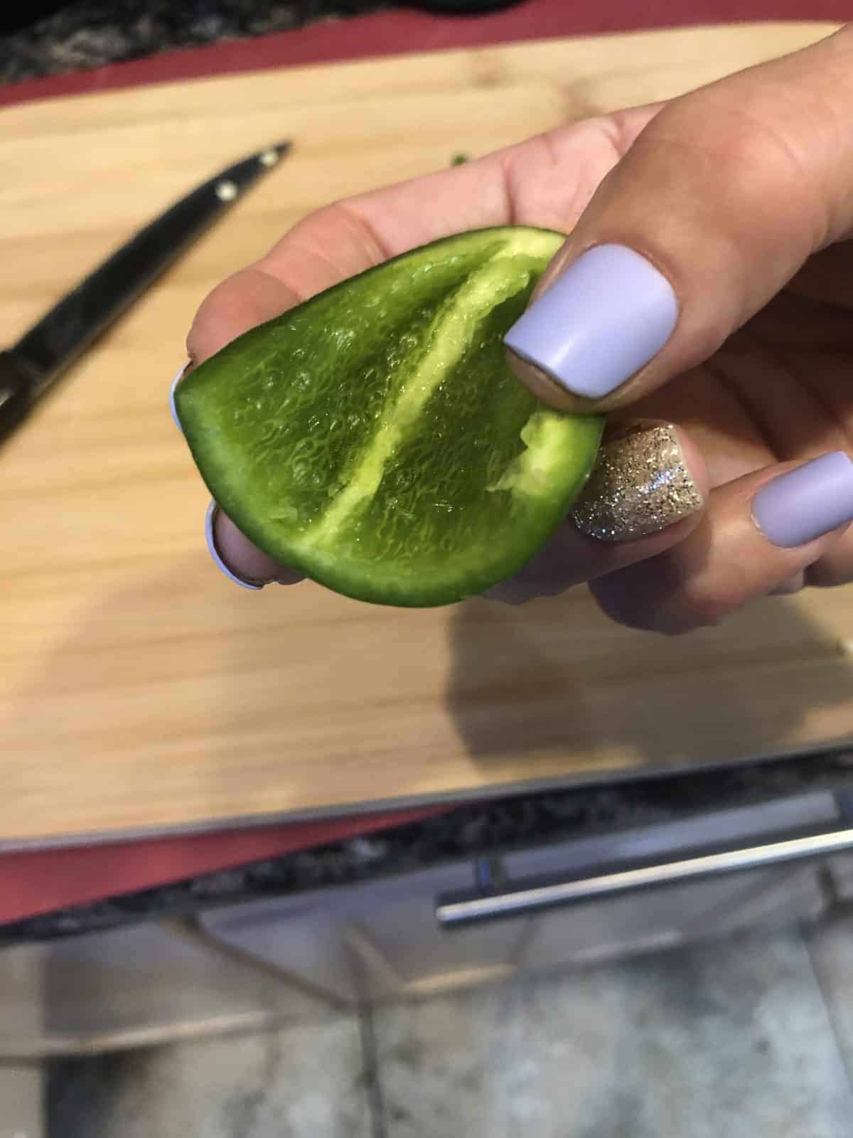 A hollowed out slice of a Jalapeno being held by a hand with purple nail polish over a wooden cutting board with a knife on the side