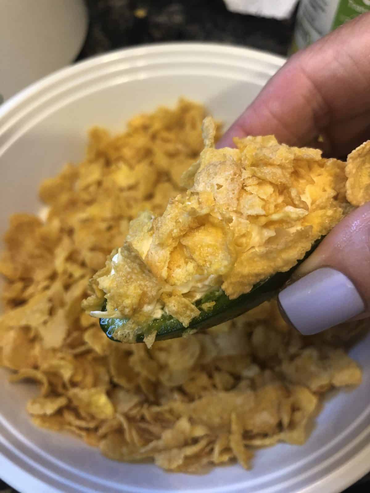 Cornflake Breaded Jalapeno Poppers being held in a hand with purple nail polish on nails over a bowl of corn flakes