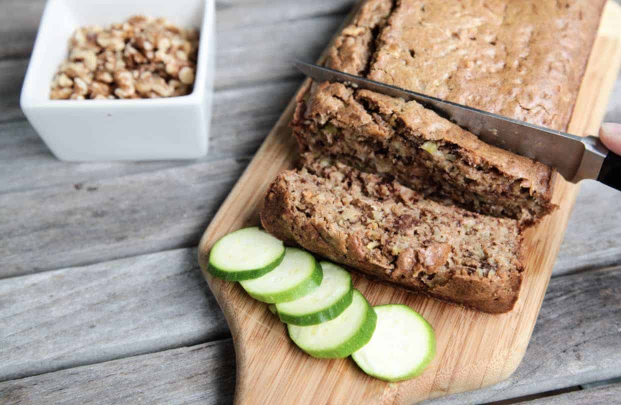 A loaf of sliced Low Carb Zucchini Bread being cut with a knife on a wooden cutting board with sliced zucchini pieces on the side sitting on a wooden table with a small white bowl filled with oats