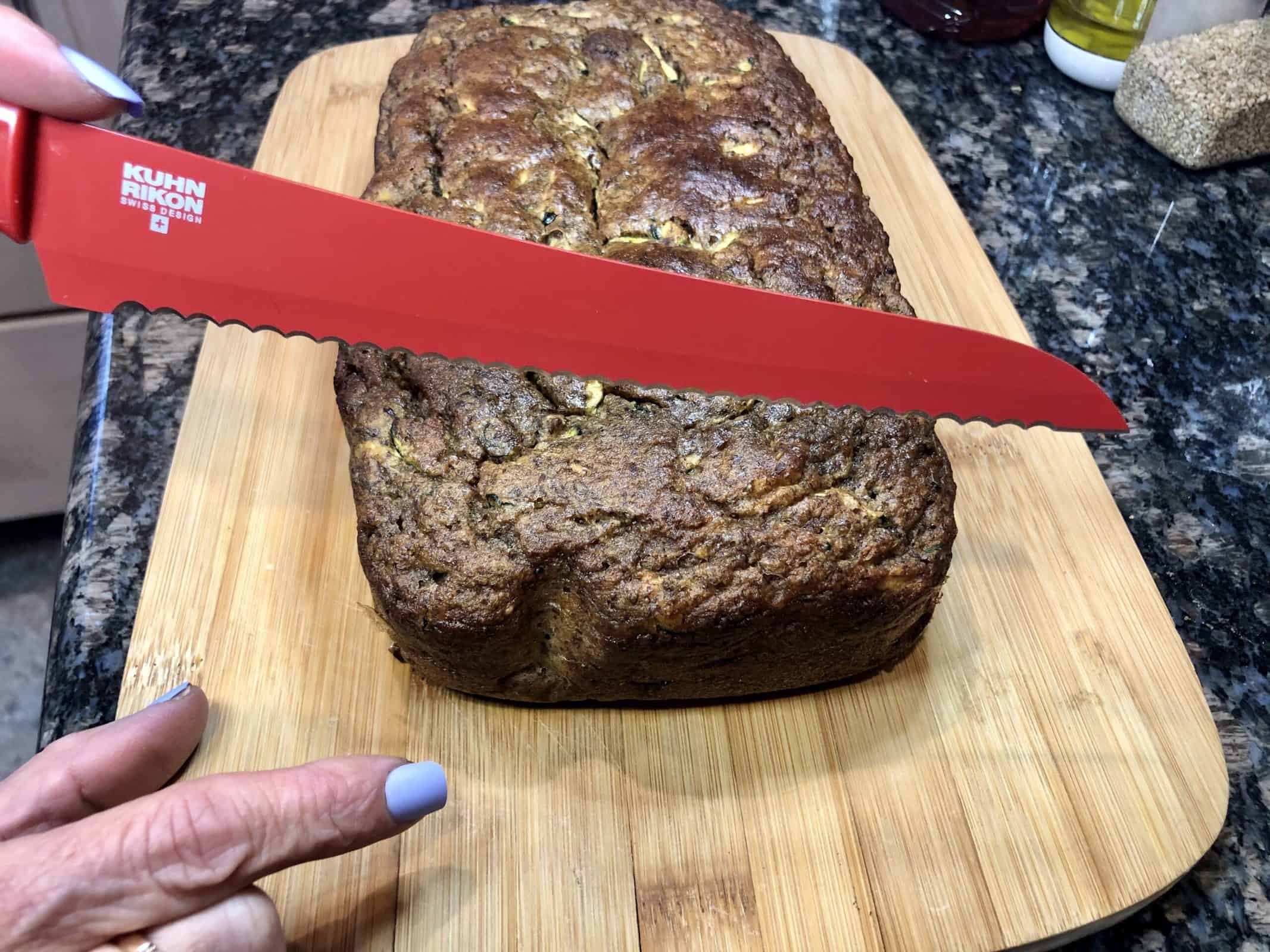 A loaf of Homemade Zucchini Bread on a wooden cutting board about to be sliced with a red knife