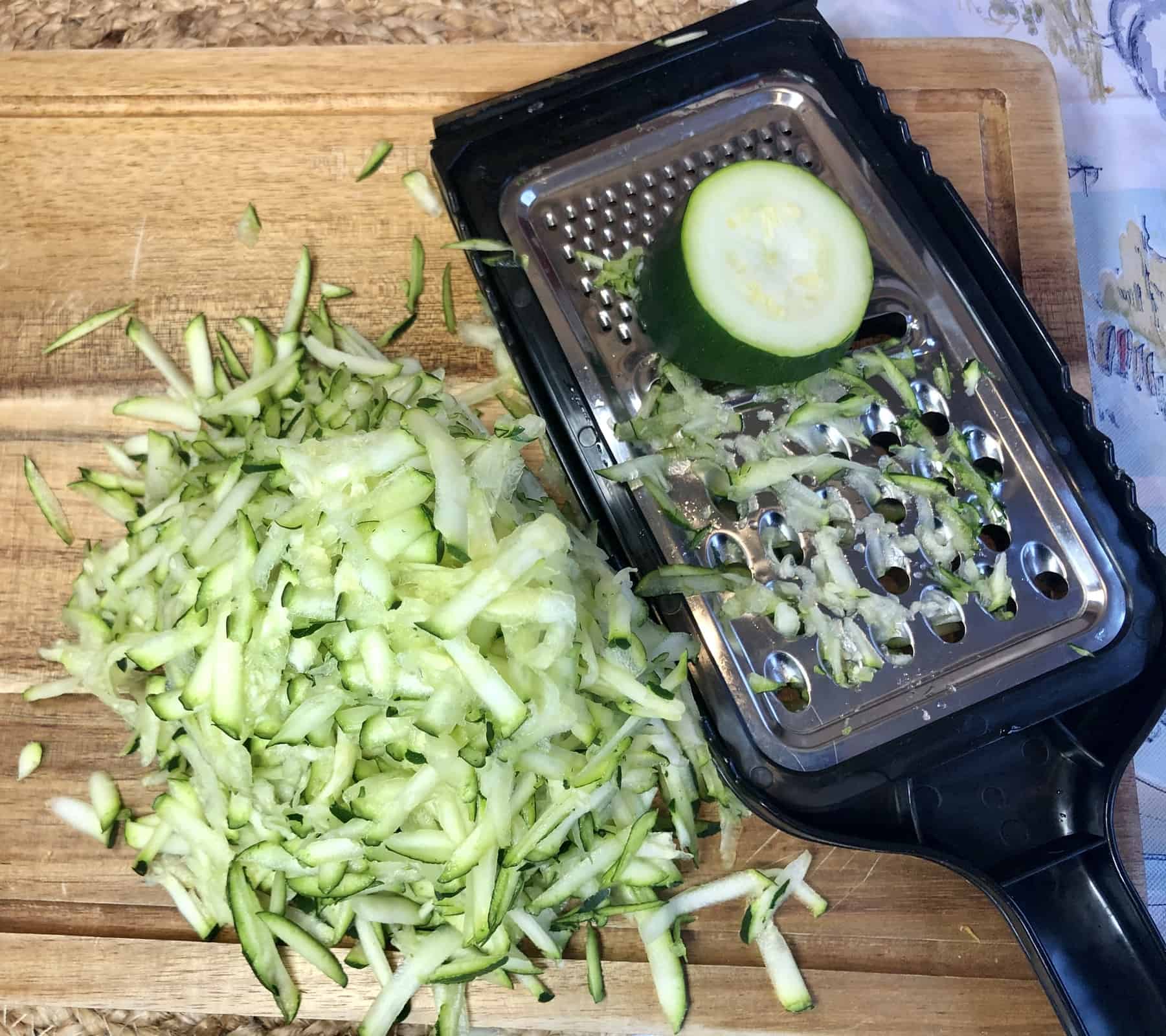 Grated Zucchini on a wooden cutting board with a black grater and zucchini on the side