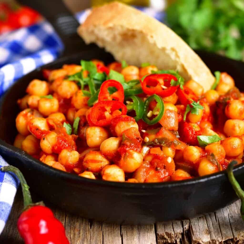 Spiced chickpea stew in a black bowl with a slice of Italian bread dipped in the bowl on a wooden table with a blue checkered napkin