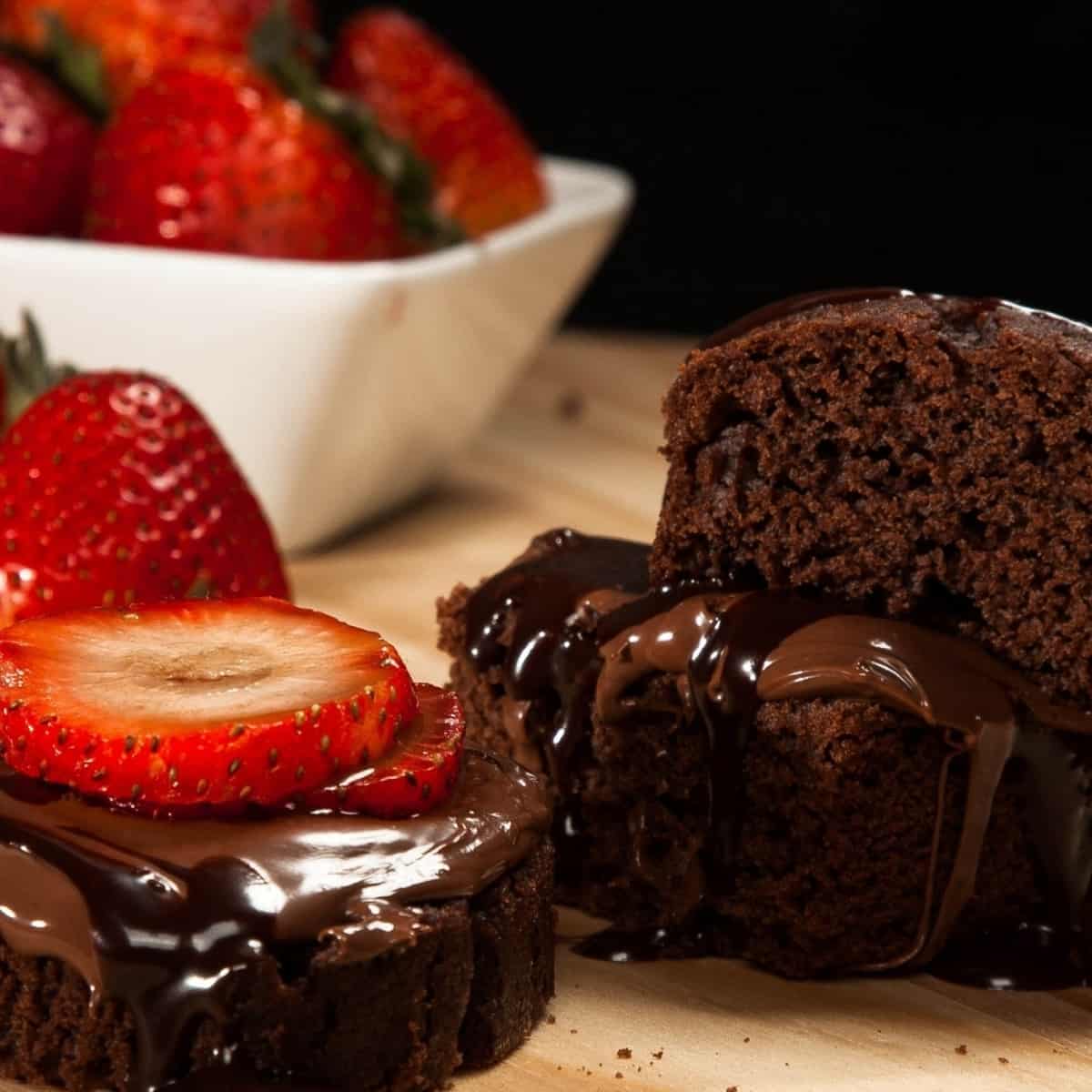 Sliced Chocolate Cake drizzled with chocolate syrup on a wooden cutting board with Strawberries and a white bowl holding strawberries