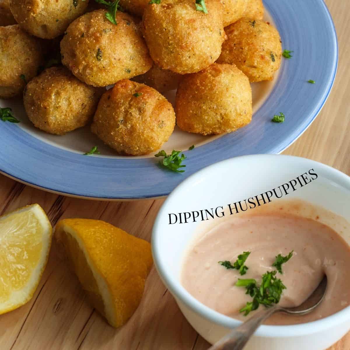 A stack of Hushpuppies on a blue dinner plate on a wooden cutting board with a side of Aioli Sauce in a small white bowl with sliced lemons on the side