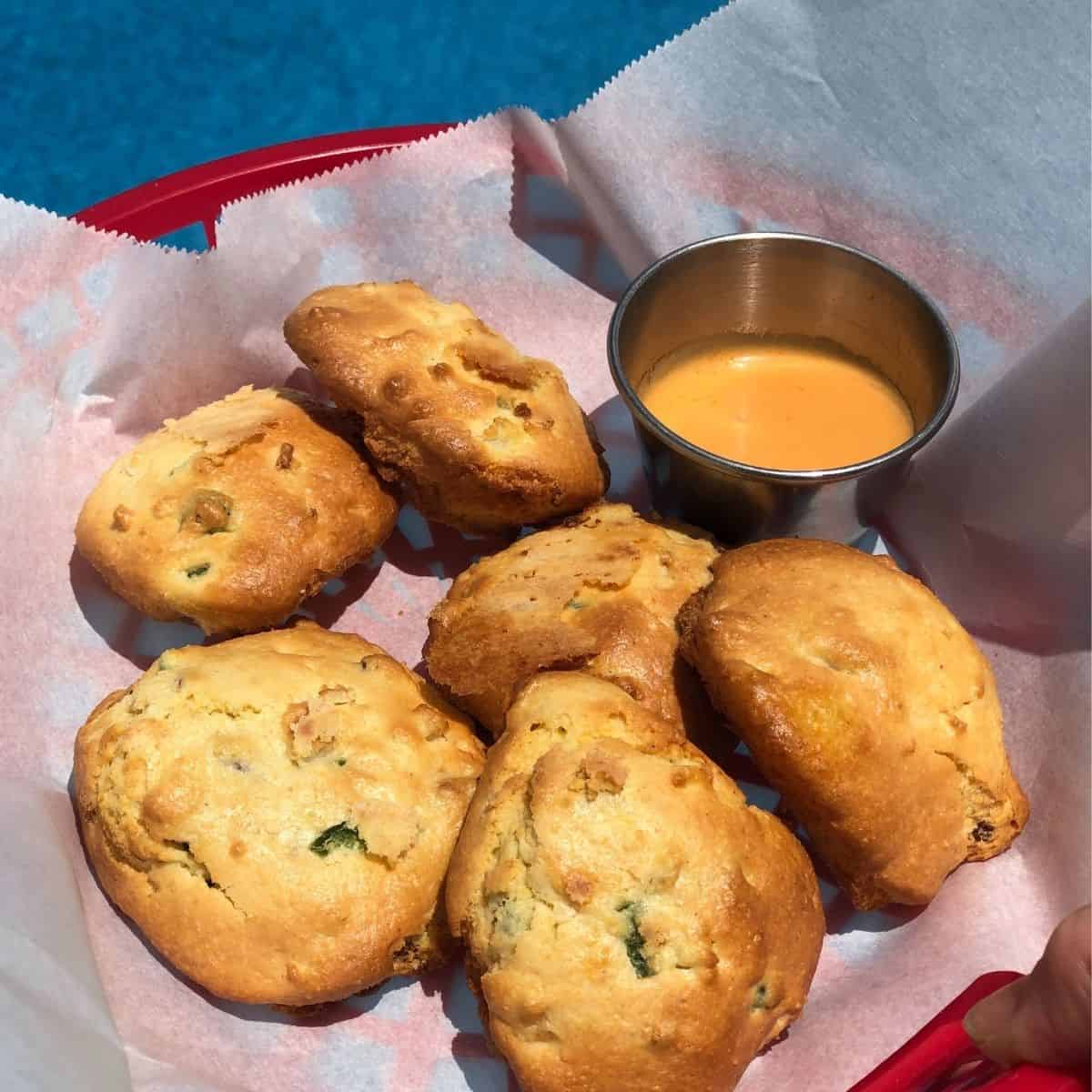 Hushpuppies in a red basket on top of parchment paper with a stainless steel cup with dipping sauce situated next to a pool