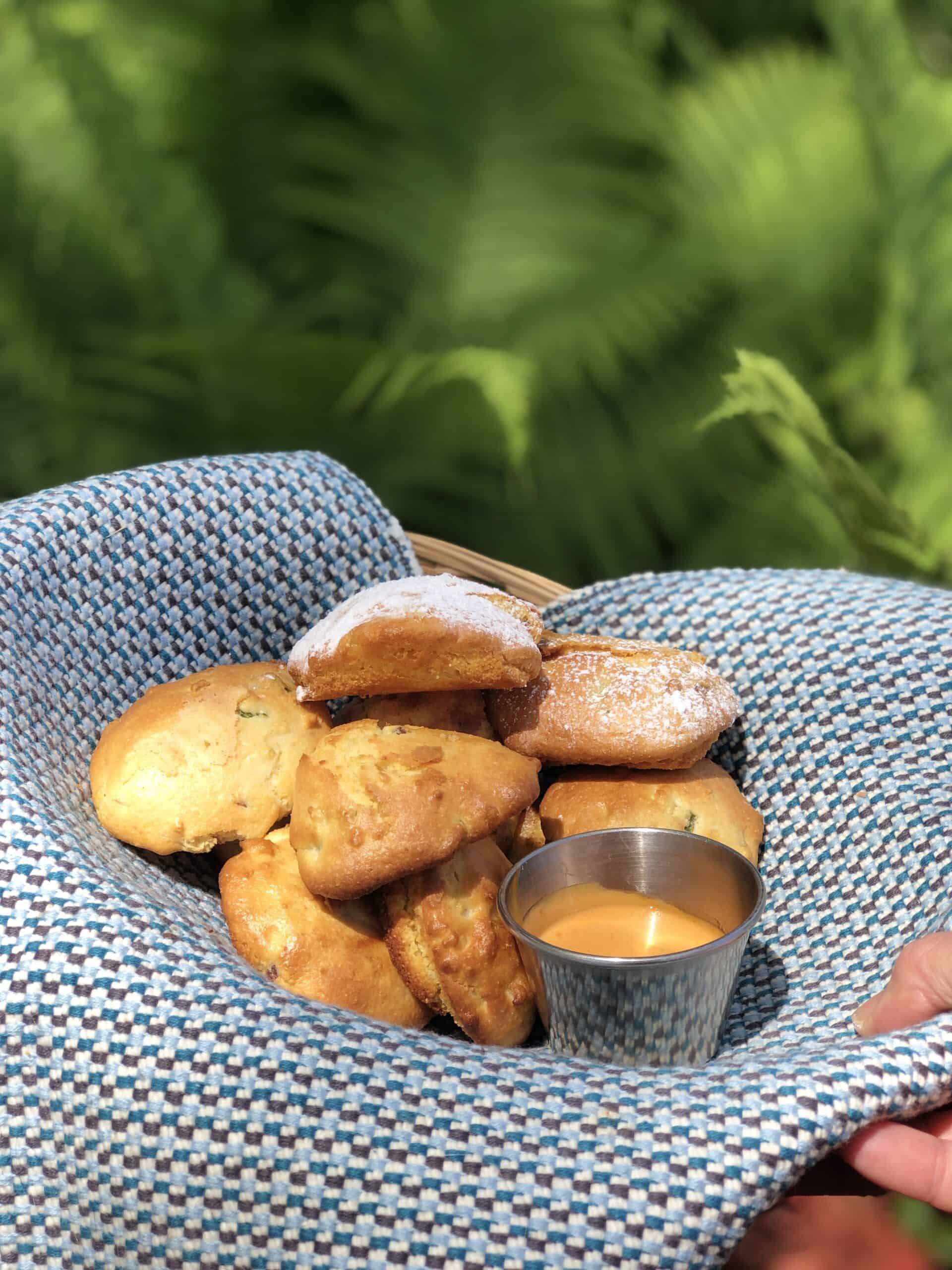 Basket of Southern Hushpuppies being held by a hand on a blue fabric placemat with a side of dipping sauce in a small stainless steel vessel