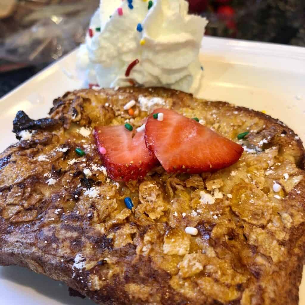Keto French Toast Crunch slice with strawberries on top and colored sprinkles and whipped cream on a white plate