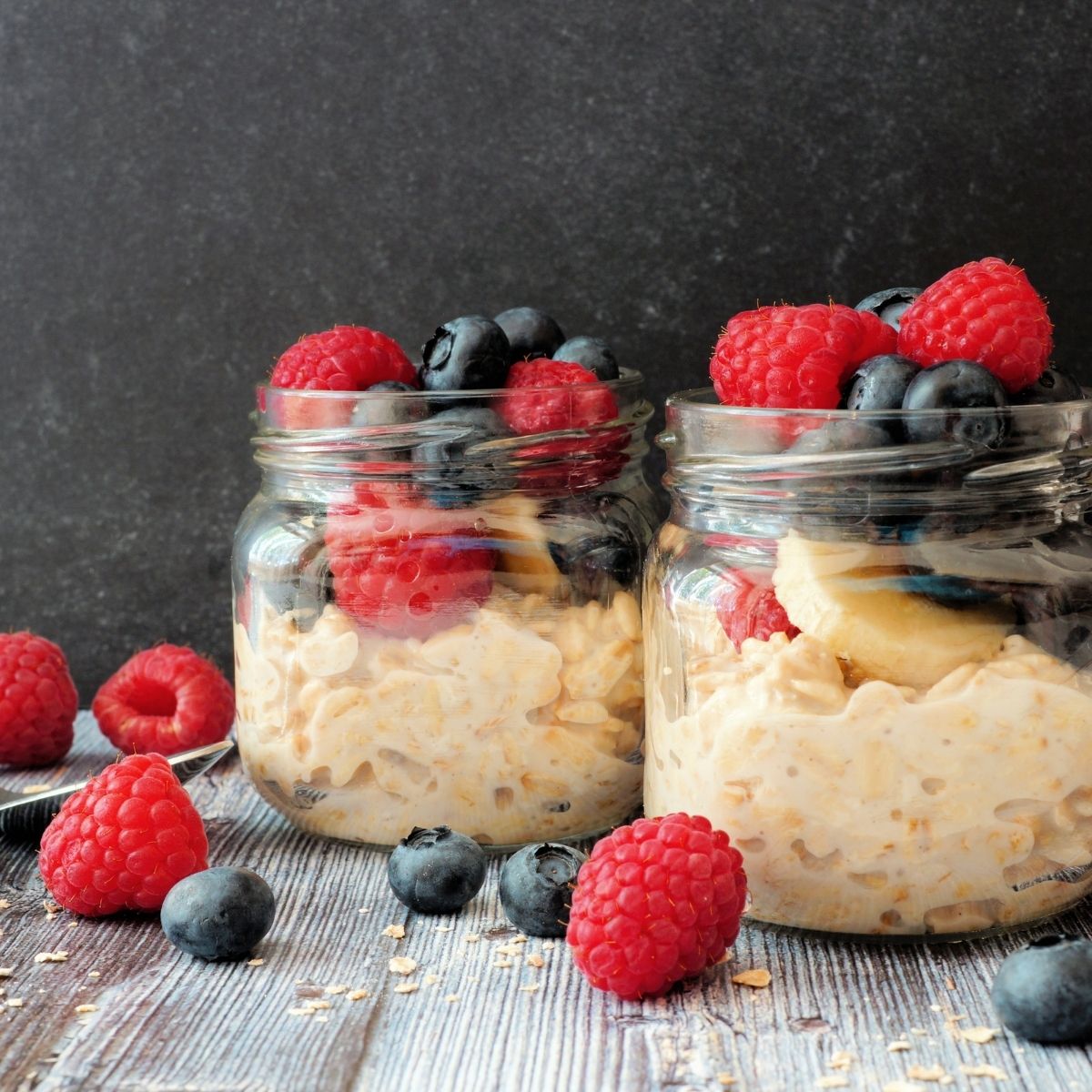 2 clear glass mason jars filled with overnight oats with raspberries, blueberries and sliced bananas on a wooden table covered in raspberries and blueberries