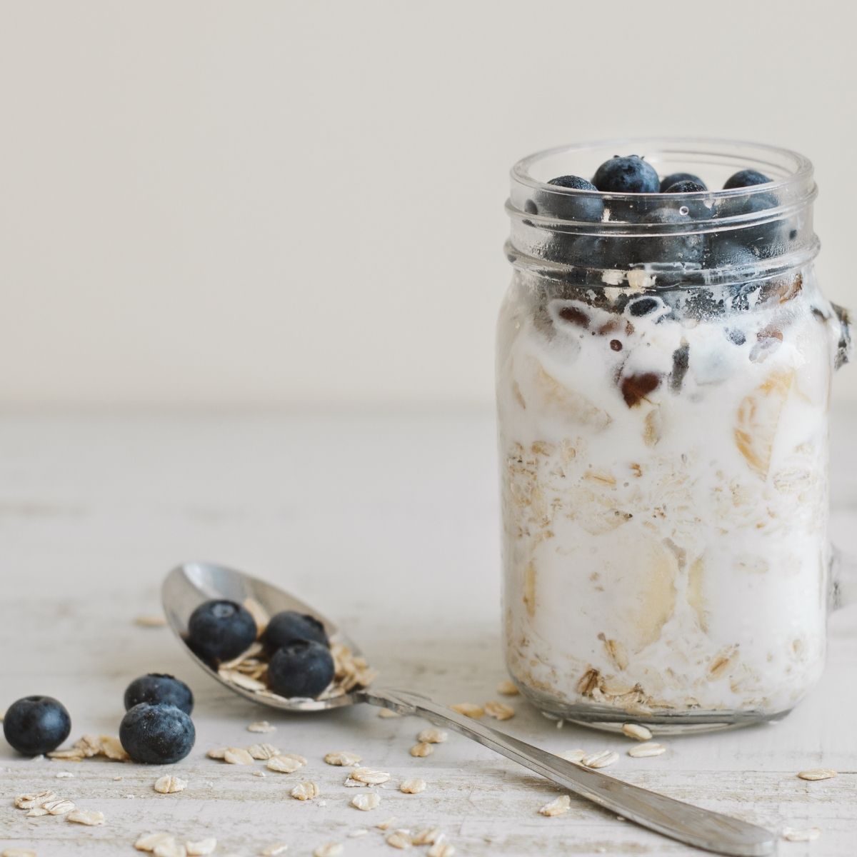Overnight Oats with blueberries on top in a clear mason jar mug sitting on a table with a spoon holding two blueberries and 2 blueberries sprinkled on the table with oats