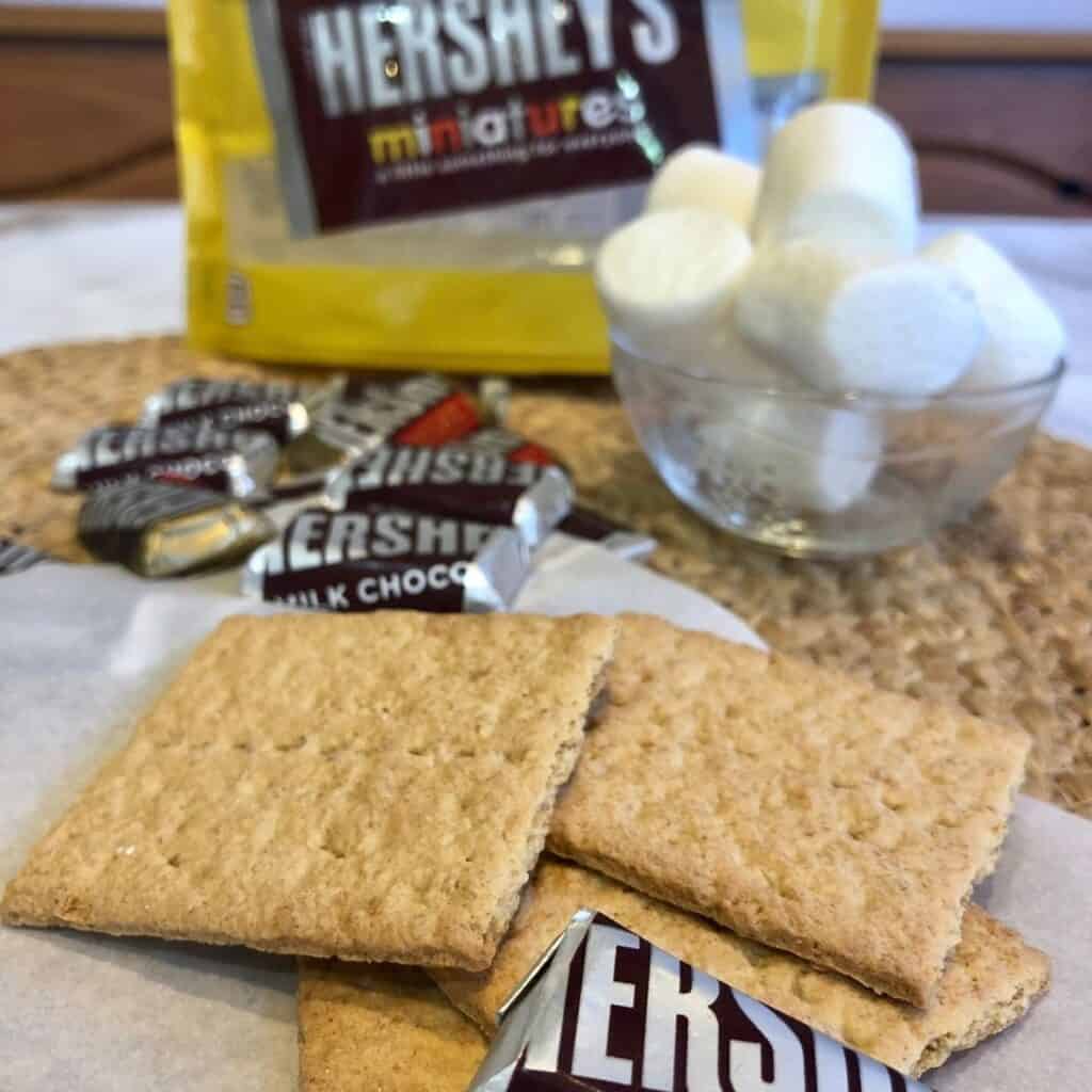 Graham crackers, miniature Hershey bars and marshmallows in a clear glass bowl on a table with a bag of Hershey's chocolates