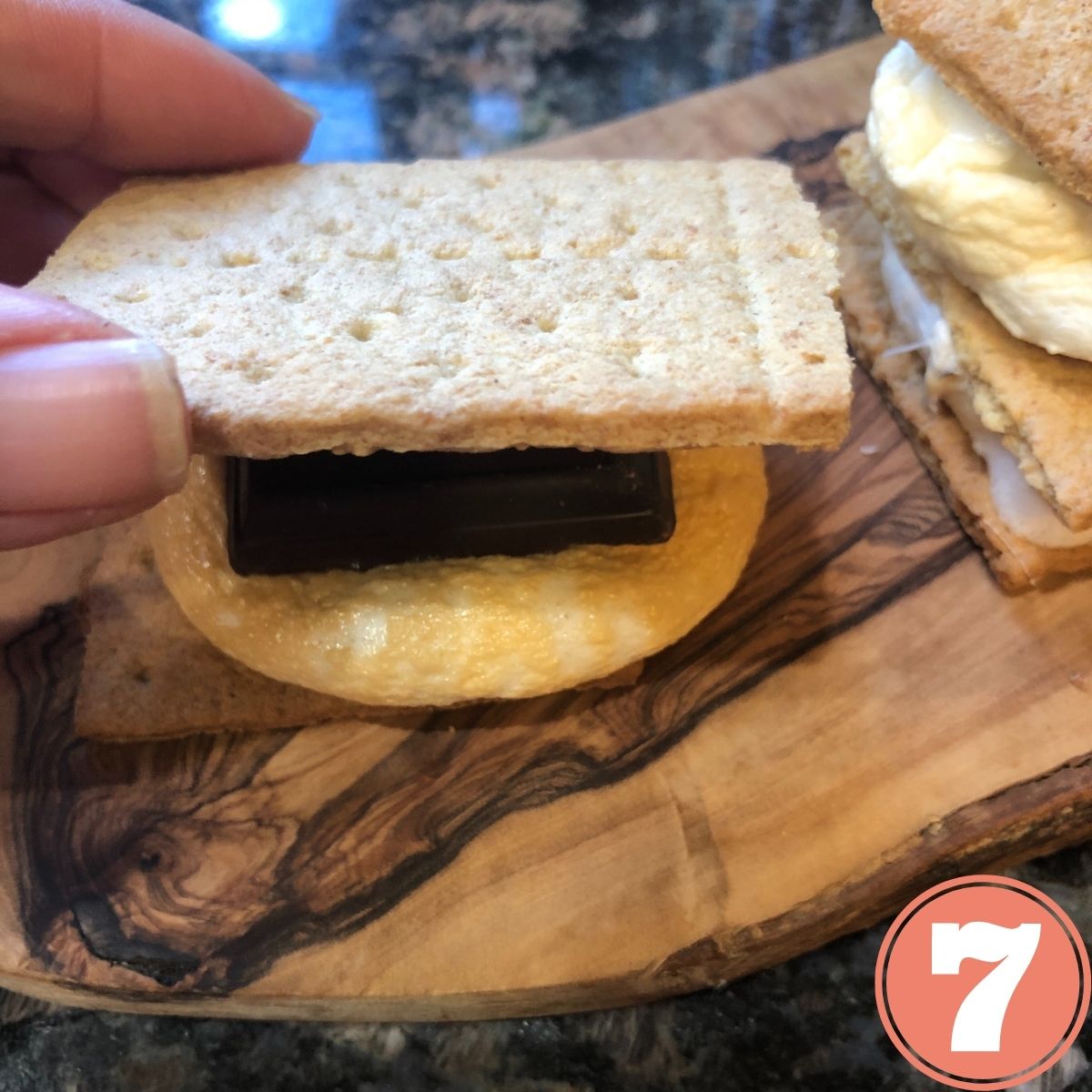 A hand placing a graham cracker on top of a chocolate bar sitting on a toasted marshmallow