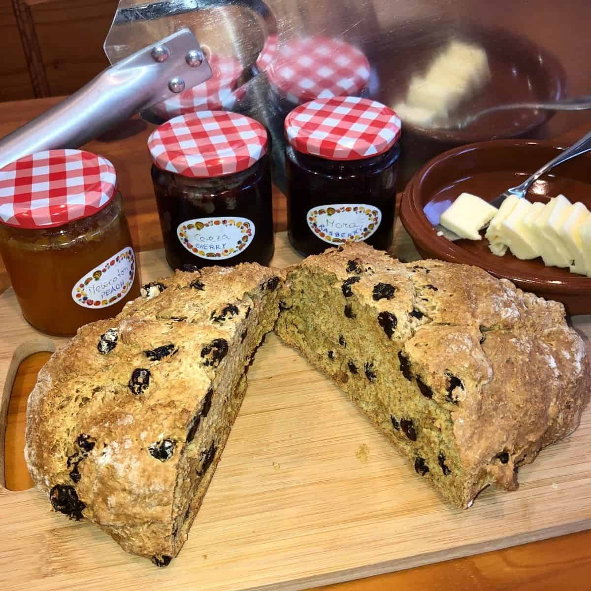 Irish Soda Bread with Raisins sliced in half on a wooden cutting board with 3  jars of peach jam, cherry jam and raspberry jam and a brown bowl with sliced butter