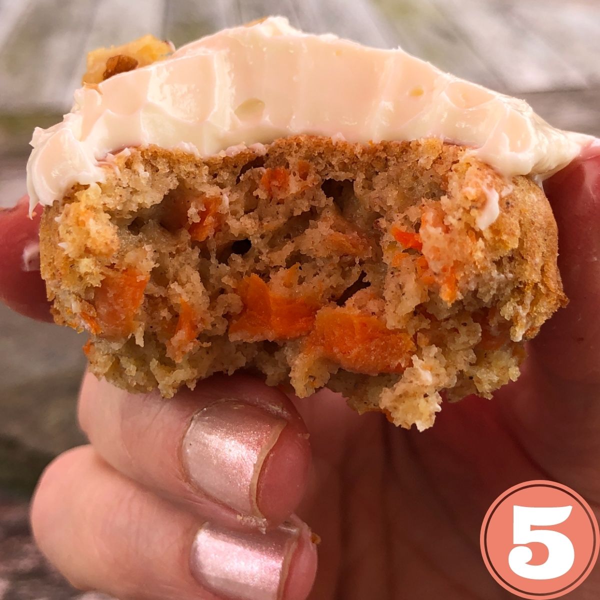 A hand holding a bite of a  cream cheese iced carrot cake muffin
