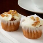 Two carrot cake muffins with cream cheese frosting and walnuts on top sitting on a white plate