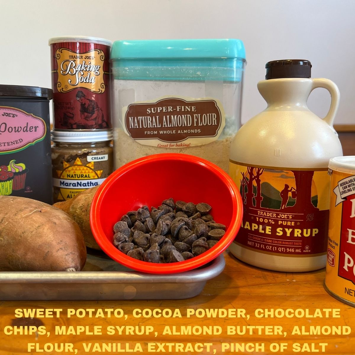 Triple chocolate brownie ingredients on a wooden table - cocoa powder, almond flour, almond butter, sweet potatoes, chocolate chips in a red bowl and maple syrup